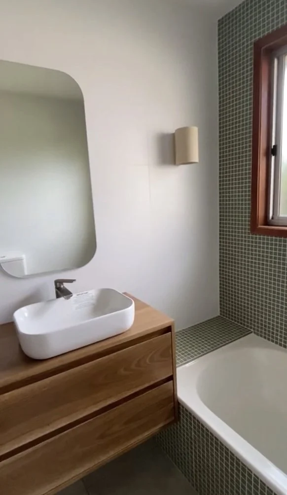 Modern bathroom with wooden vanity, white vessel sink, wall-mounted mirror, small green tiled shower area, and a window with wooden trim.