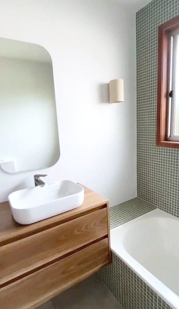 Modern bathroom with a white vessel sink on a wooden vanity, a large mirror, a wall-mounted light fixture, a window with wood trim, and a tiled bathtub surround.