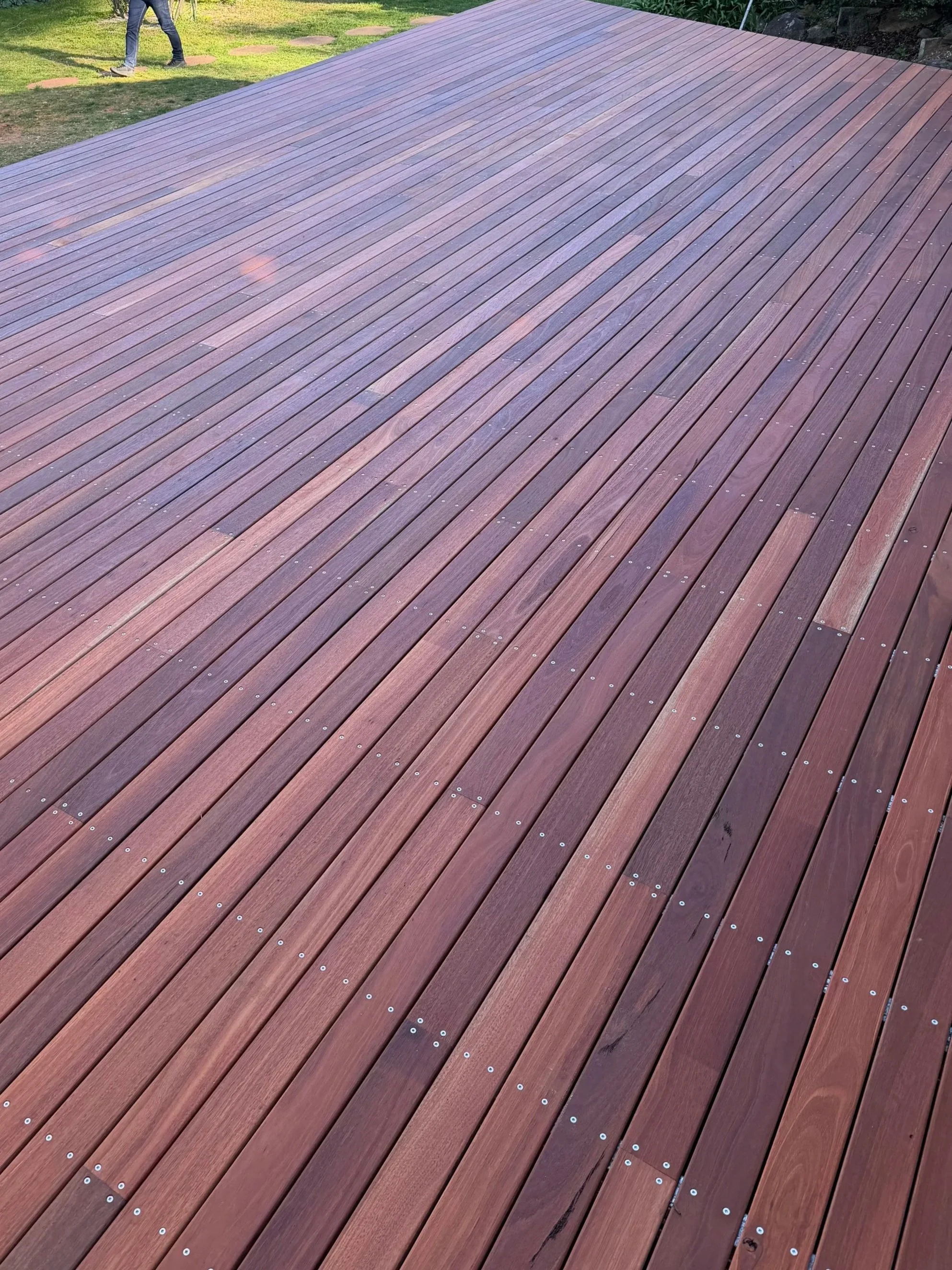 A wooden deck with curved planks and small white screws, adjacent to a grassy yard where a person is walking on stepping stones.