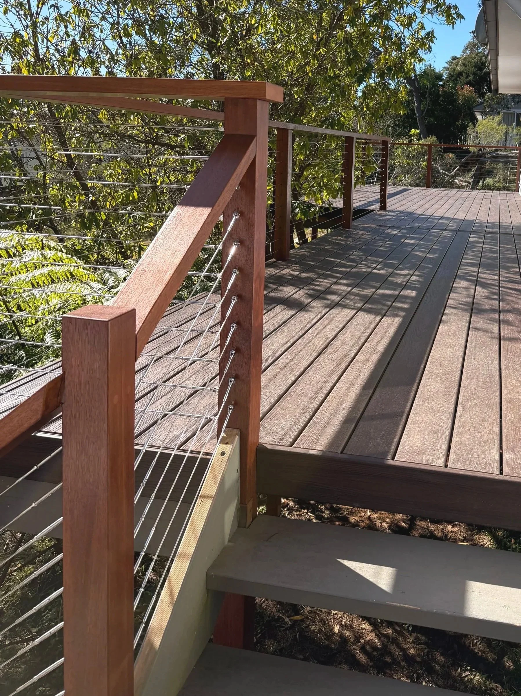 Wooden deck with railing and wire cable fencing, surrounded by trees and bushes, under a blue sky.