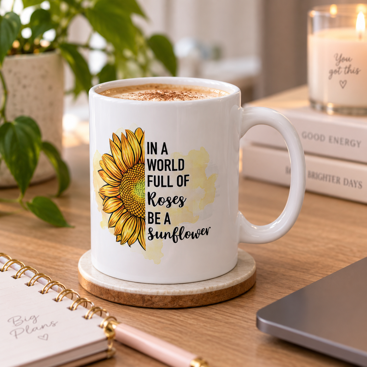 White mug with a sunflower illustration and the quote, 'In a world full of roses, be a sunflower,' on a wooden desk. There is a potted plant, a notebook, a pen, and a candle in the background.