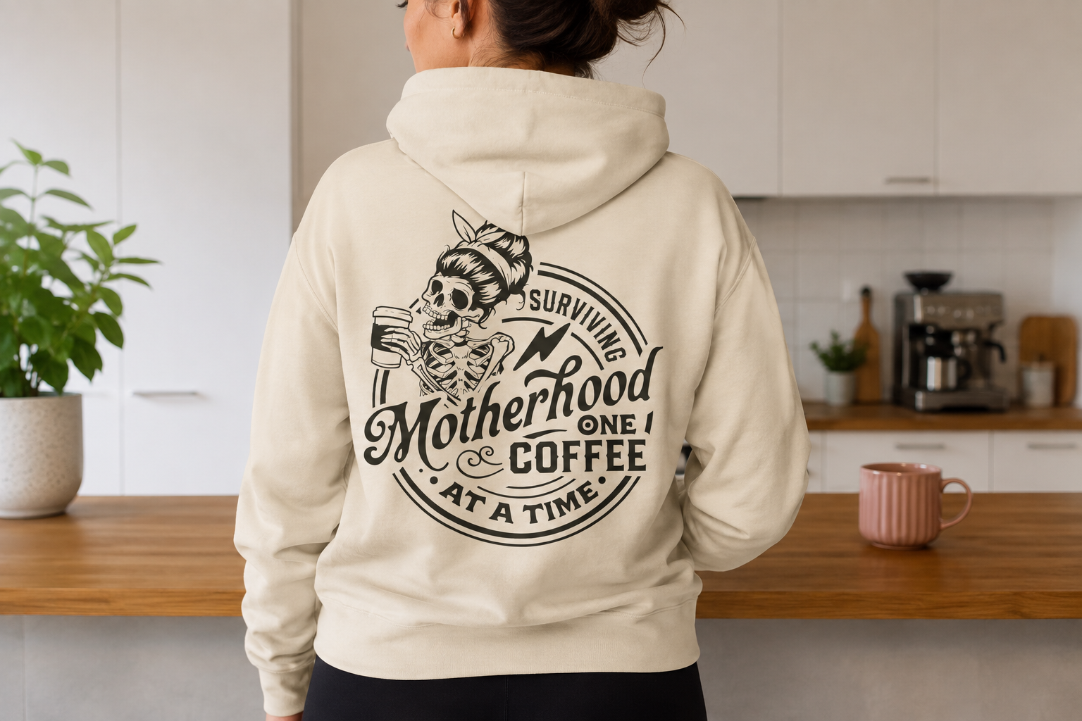 Back view of a woman in a beige hoodie with a skeletal woman graphic and text about motherhood and coffee. She is standing in a kitchen with a wooden countertop, a pink mug, and kitchen appliances visible.