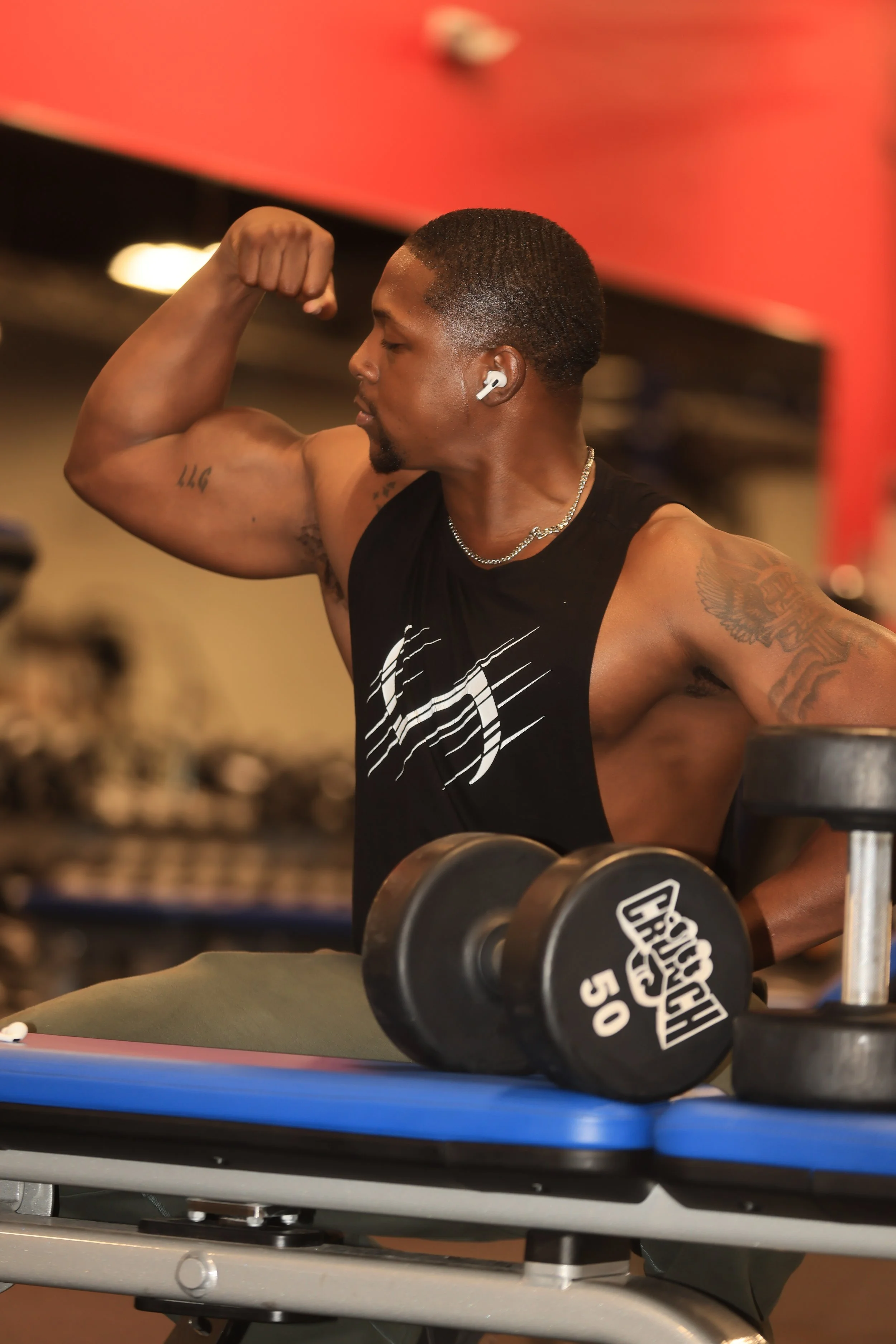 A muscular man with short dark hair and tattoos on his left arm doing a bicep flex at the gym. He is wearing wireless earbuds, a black sleeveless shirt with a white graphic, and a silver chain necklace. There are dumbbells on a blue workout bench in front of him.