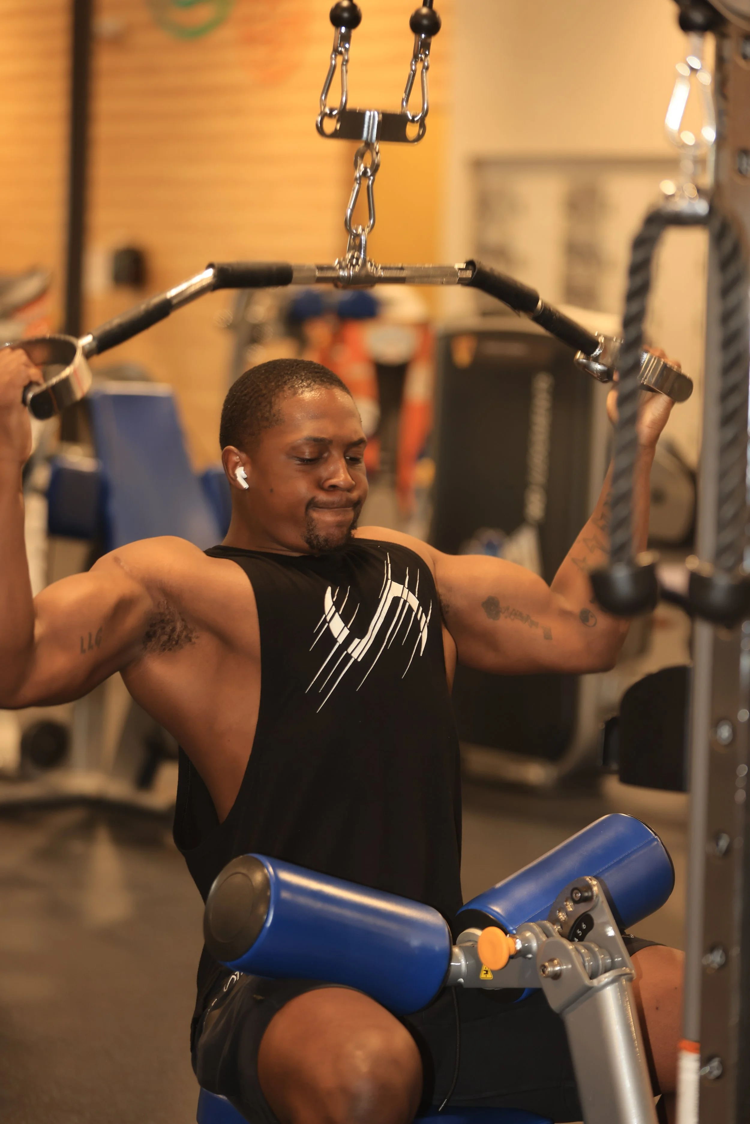 A man with short hair and earbuds working out on gym equipment, lifting weights with a focused expression.