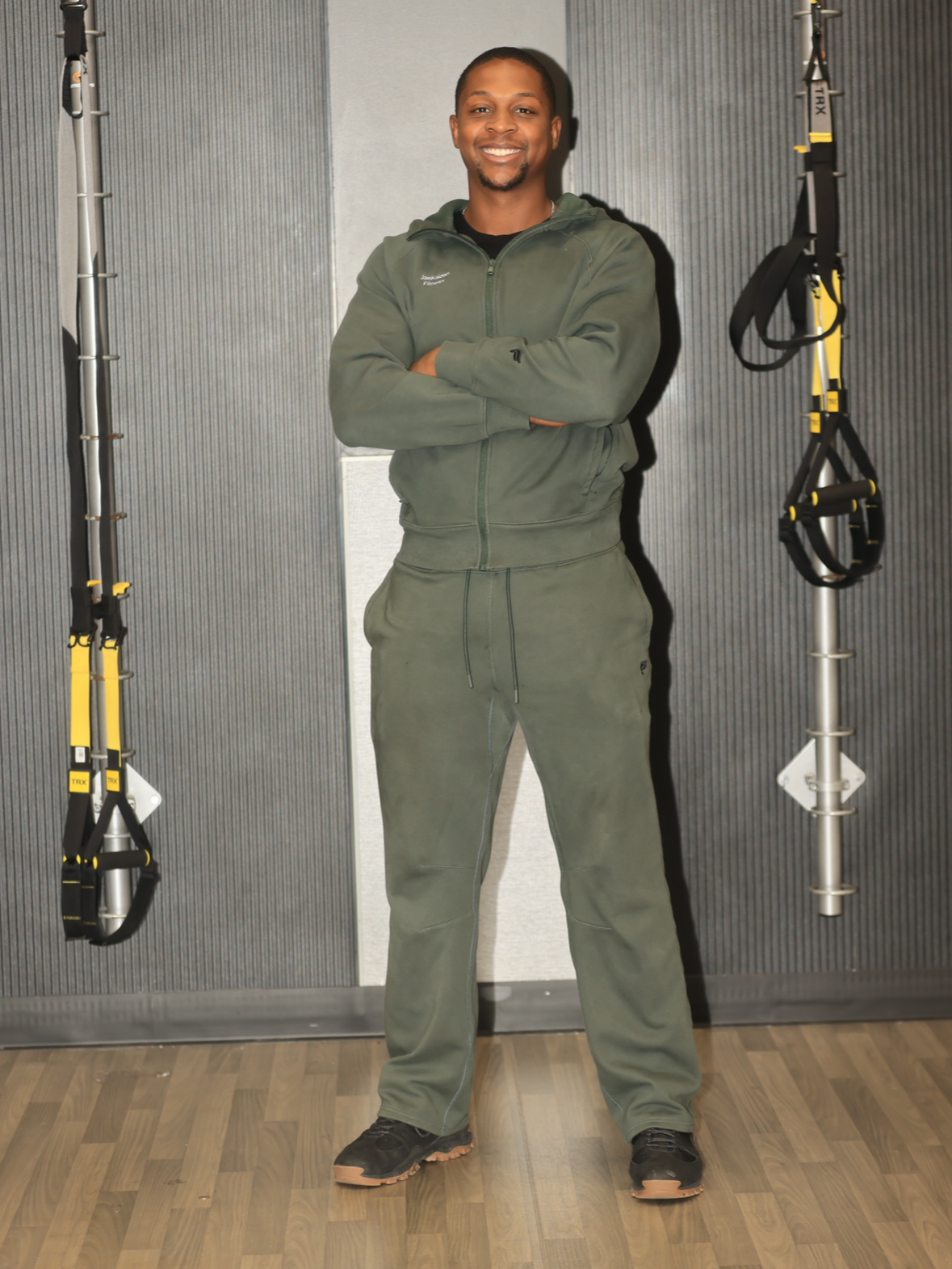 A smiling man in green athletic wear standing with arms crossed in front of a wall with suspension training equipment hanging on both sides.
