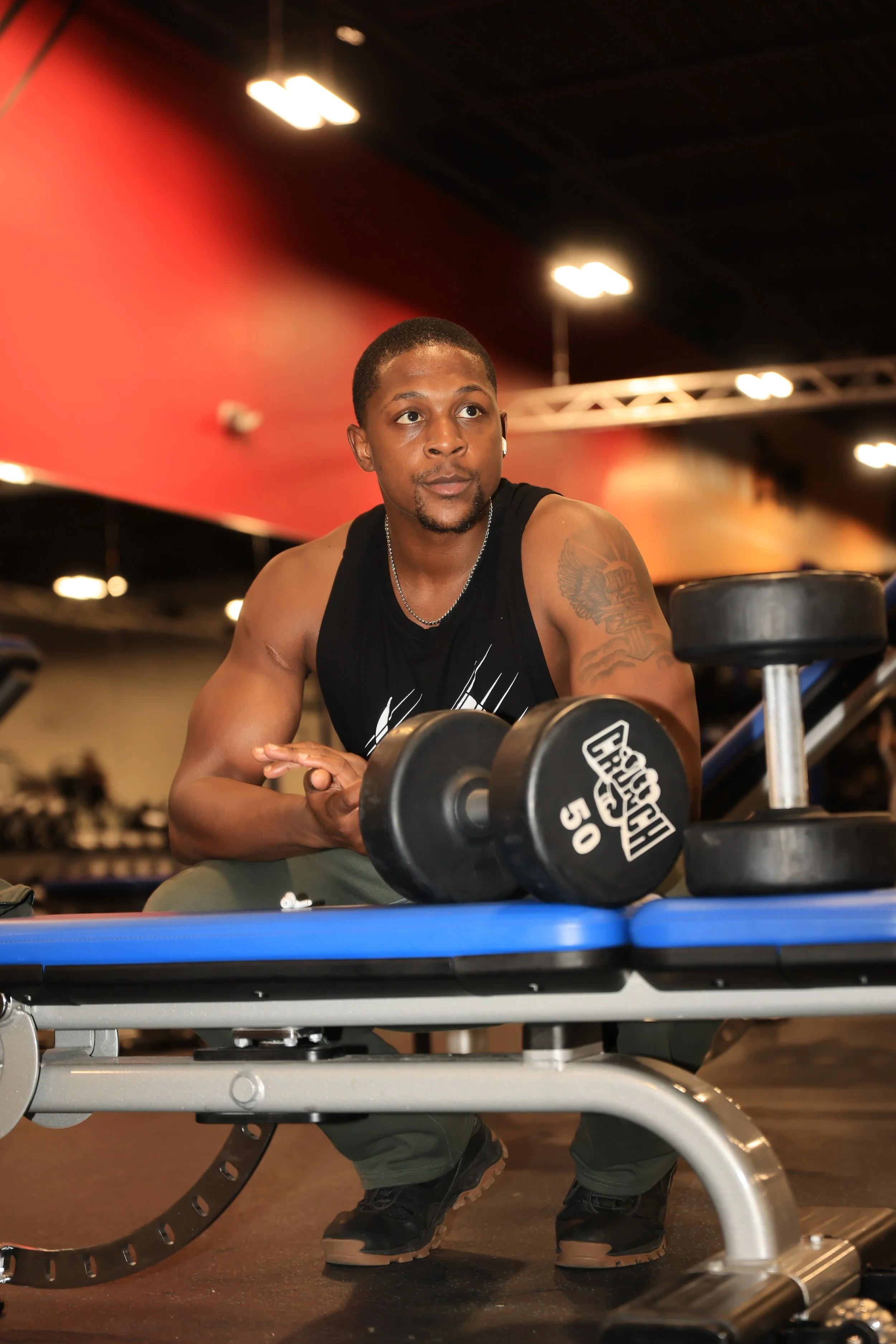 Young man with tattoos on his arm, wearing a black tank top and wireless earbuds, sitting in a gym near a table with dumbbells, with gym equipment and red walls in the background.