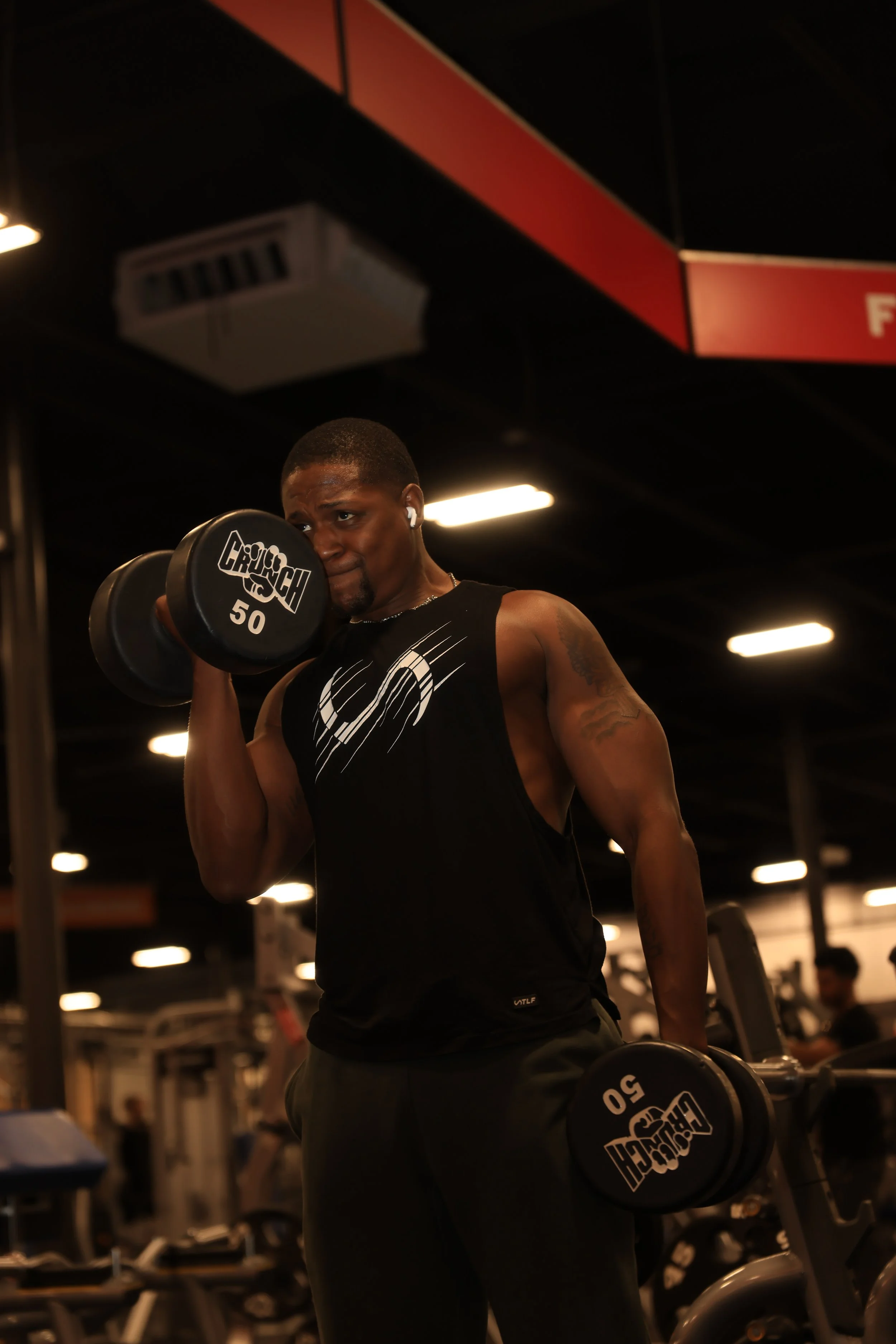 A man lifts a dumbbell in a gym, wearing a black sleeveless shirt and wireless earbuds. The gym has dim lighting with rows of exercise equipment in the background.