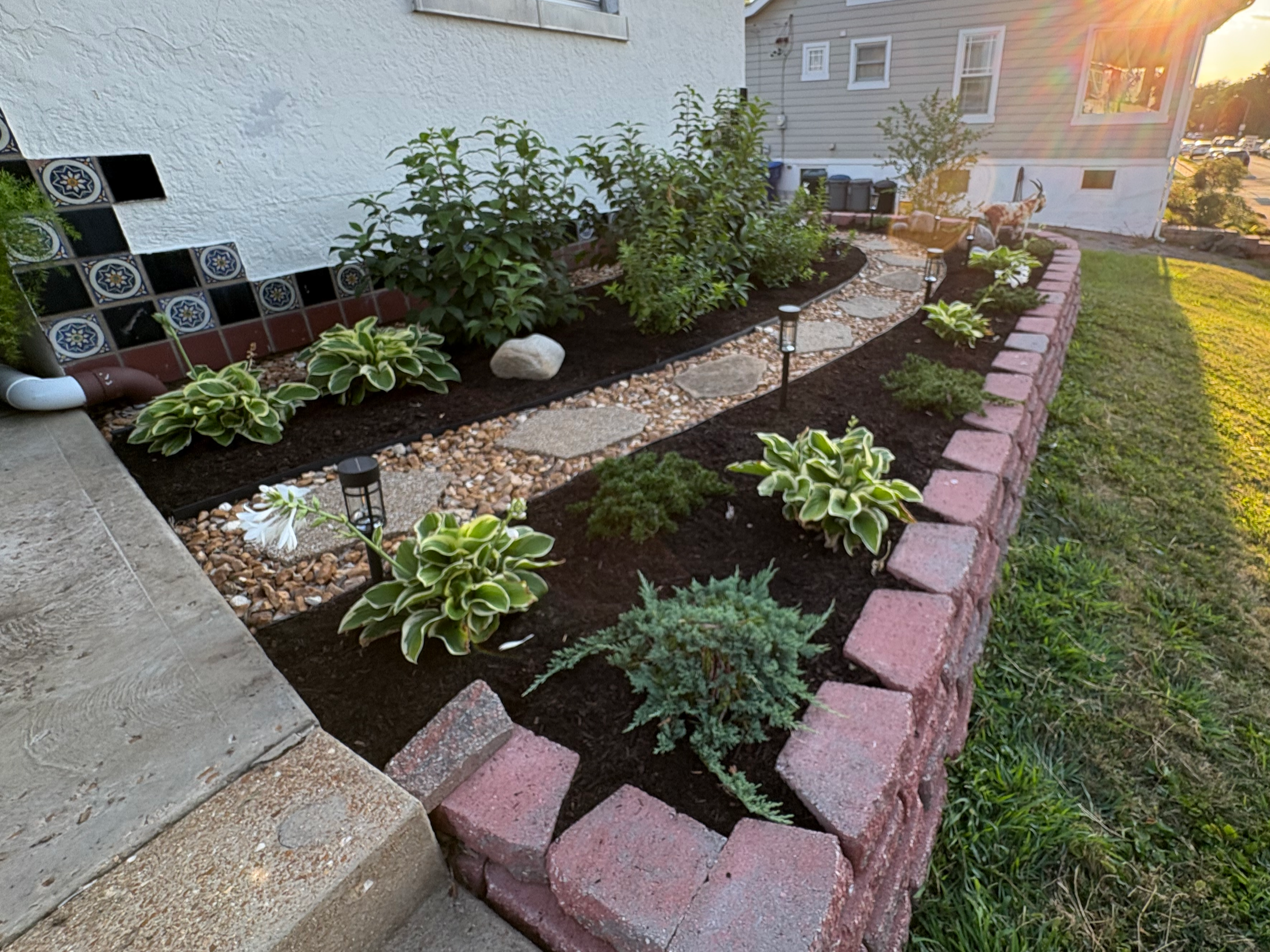 A landscaped garden bed with plants and flowers, bordered by a brick edging. A pathway of stepping stones runs through the bed, with small landscape lights along the path. Fading sunlight causes a lens flare in the background.