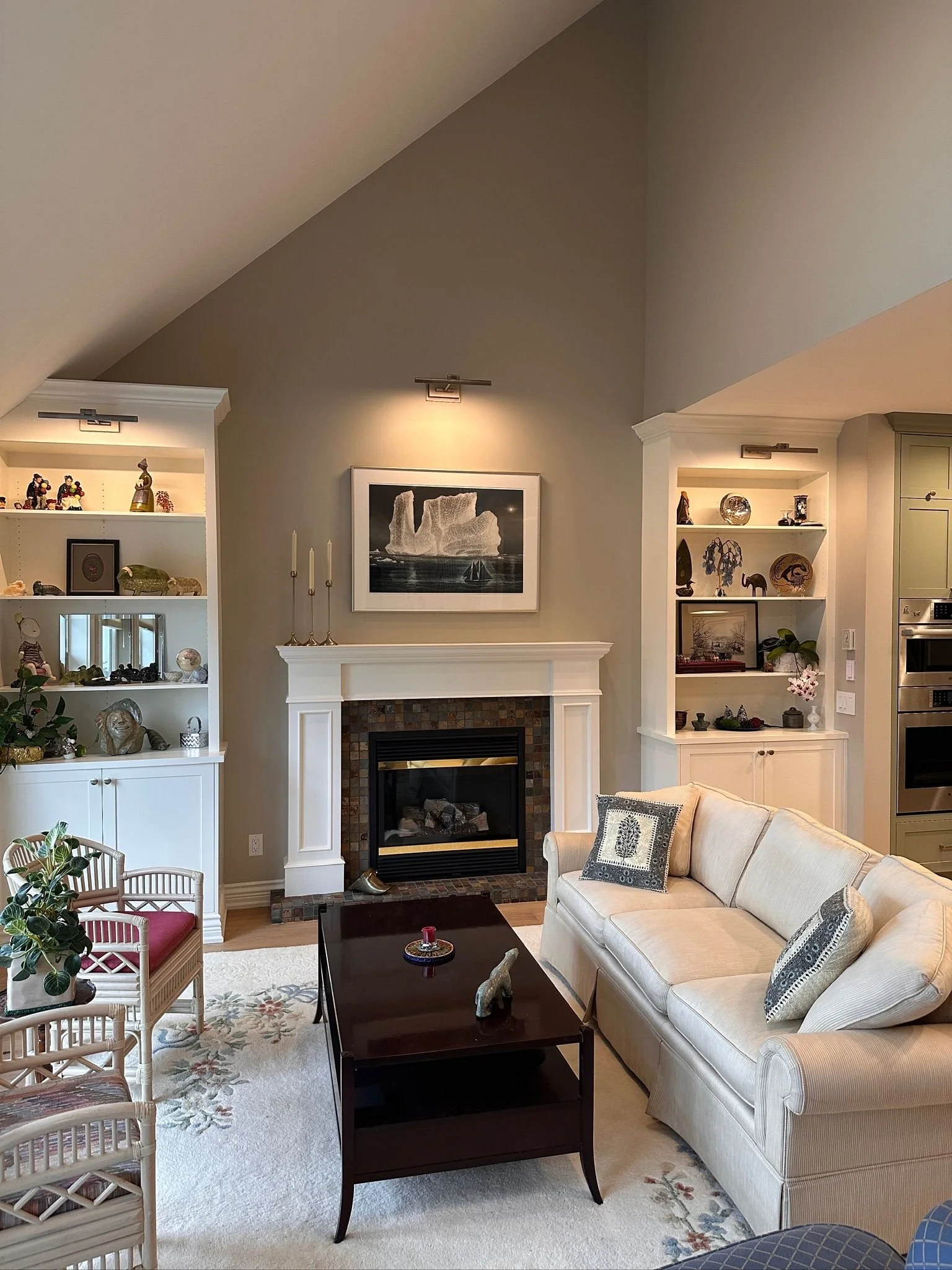 A cozy living room with a beige sofa, a black coffee table, and built-in white shelves filled with decorative items and framed pictures, centered around a fireplace with a framed artwork above it.