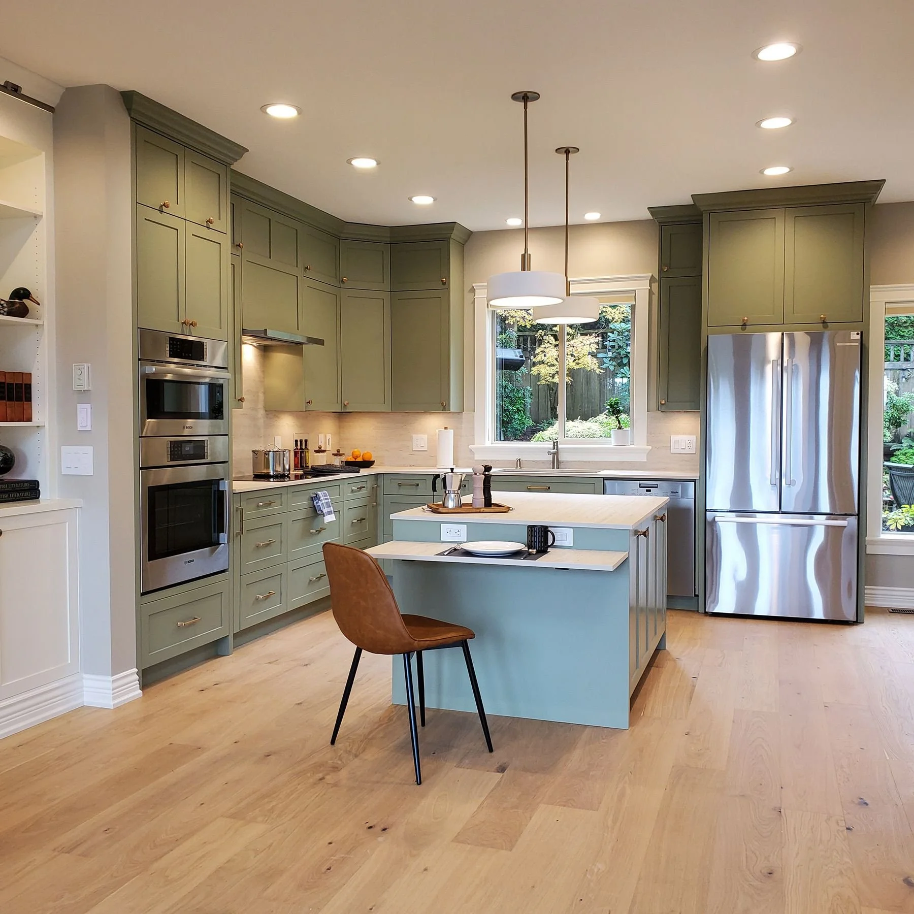 Kitchen with green cabinets, stainless steel appliances, an island with a chair, and large windows overlooking a garden.