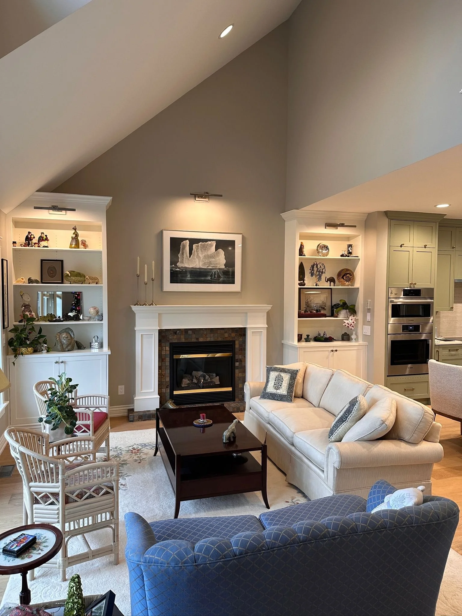 Living room with white sofa, blue armchair, fireplace, white built-in shelves with decorative items, framed art on wall, table with small objects, and kitchen area with light green cabinets.