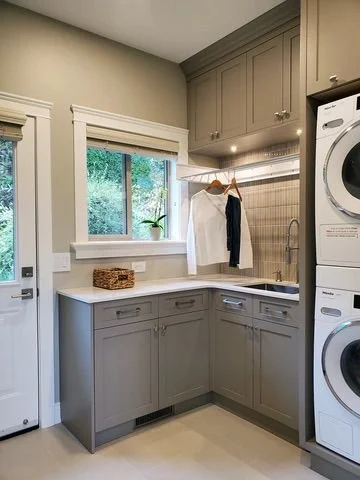 Kitchen corner with gray cabinets, a white countertop, a window with greenery outside, a drying rack with clothes, and a stacked washer and dryer.