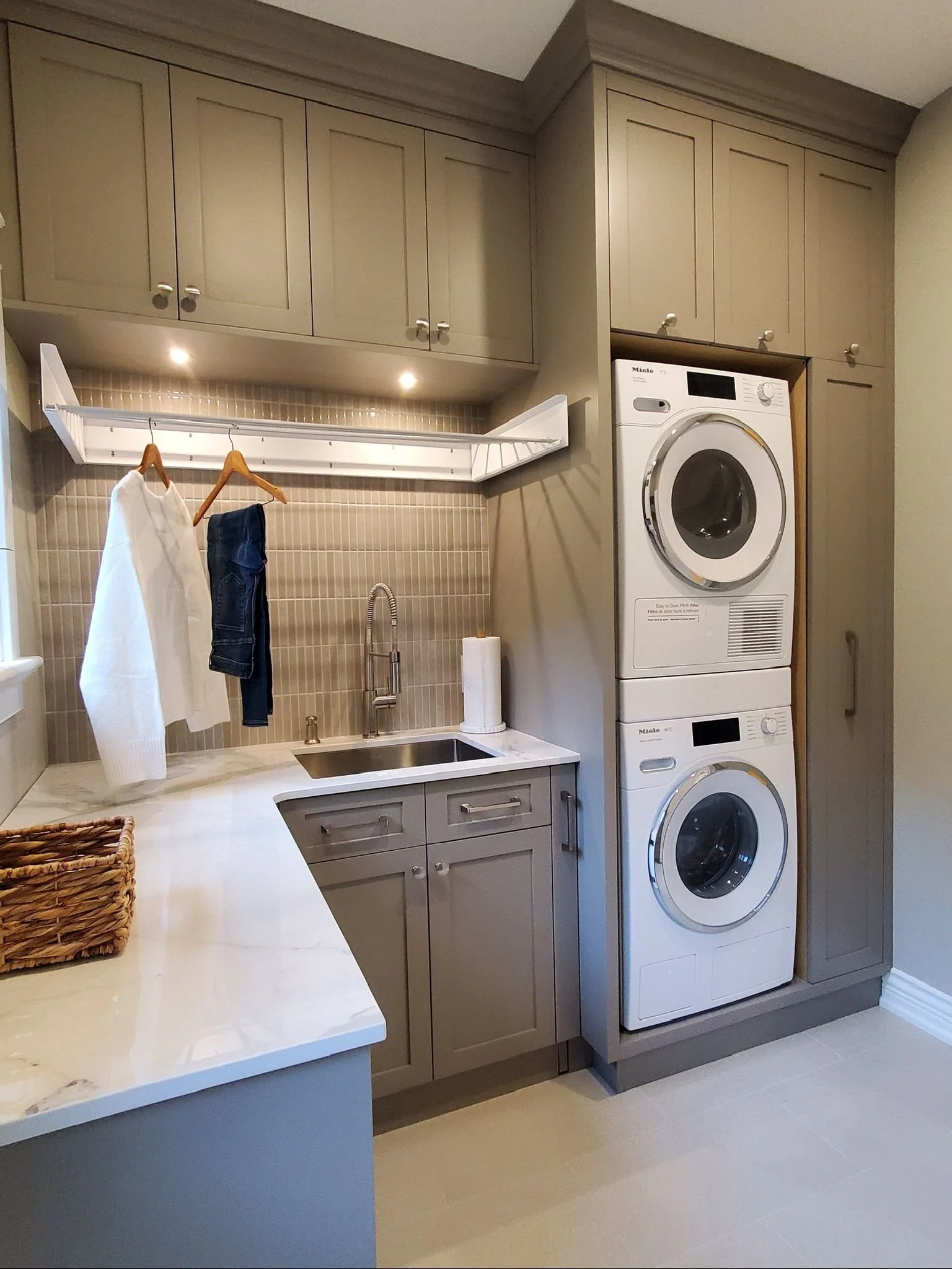 A laundry room with a washer and dryer stacked vertically on the right side. Open cabinets above the sink, hanging space with clothes, and a countertop with a wicker basket on it.