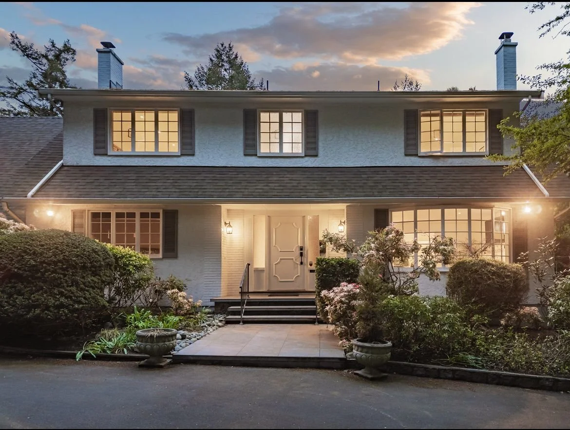 Well-lit two-story house with white exterior, dark shutters, and multiple windows, surrounded by landscaped bushes and flowers during dusk.