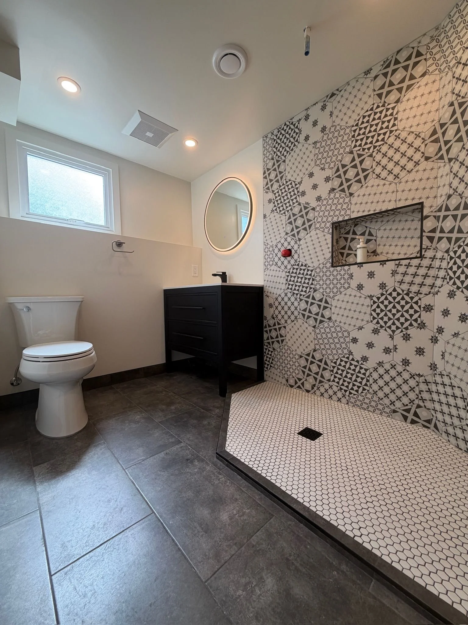 Modern bathroom with patterned tile shower area, white toilet, black vanity, circular mirror, ceiling light fixtures, window, and grey floor tiles.
