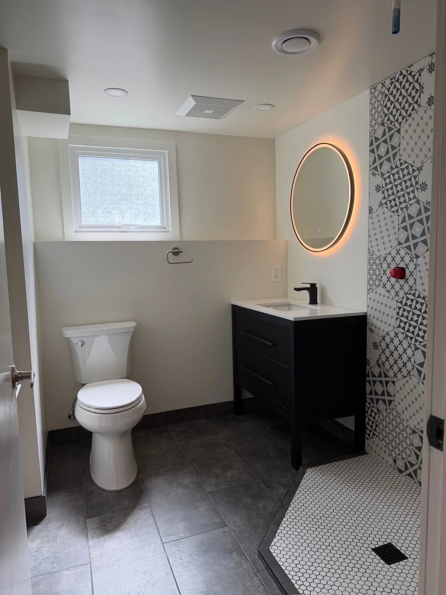 A modern bathroom with a toilet, a black vanity with a white sink, a round illuminated mirror, and a patterned walk-in shower with gray and white tiles.