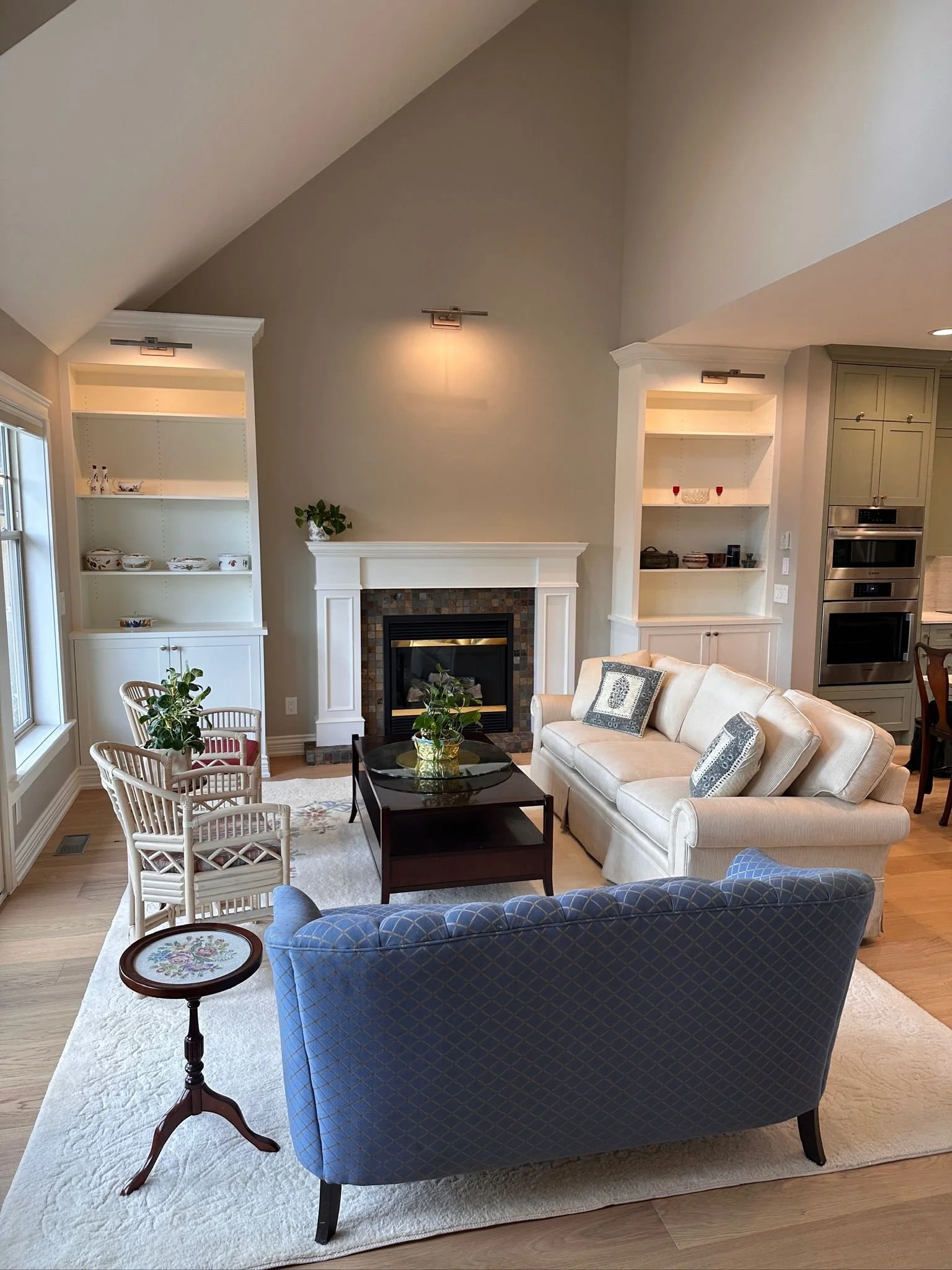Living room with a beige sofa, a blue armchair, a glass coffee table, a white rug, and a fireplace with built-in bookshelves on either side. There are plants, decorative items, and sunlight from large windows.