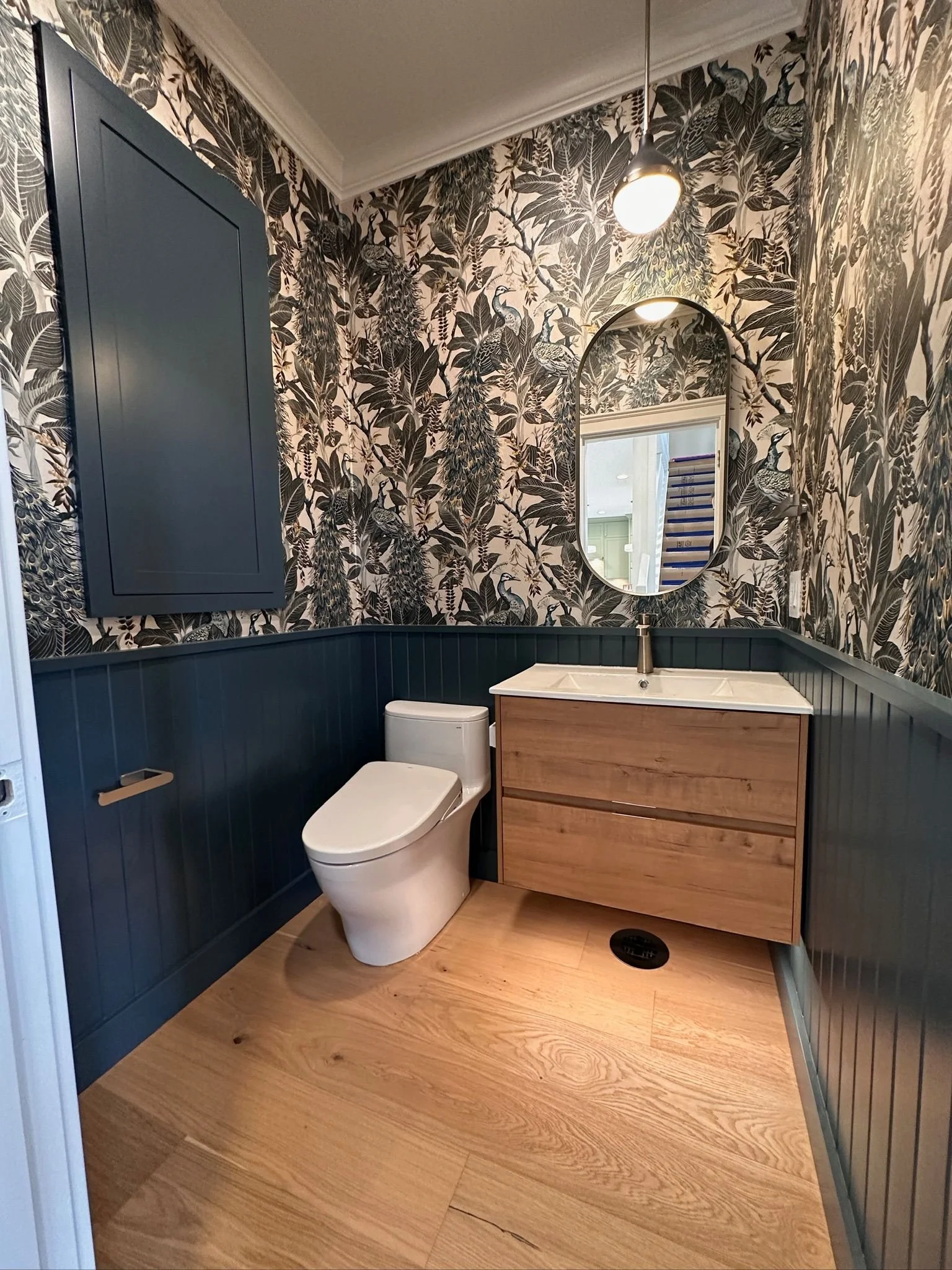 A bathroom with a peacock-themed wallpaper, a blue cabinet, a wooden vanity with a ceramic sink, a white toilet, and a large mirror. The room has wooden flooring and a pendant light fixture.