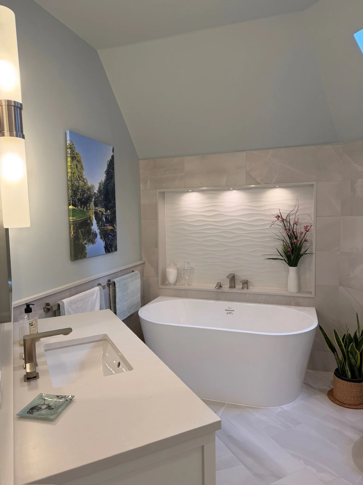Modern bathroom featuring a white bathtub, decorative wall niche with plants and lighting, bathroom vanity with sink, framed artwork of a nature scene, and potted plant.