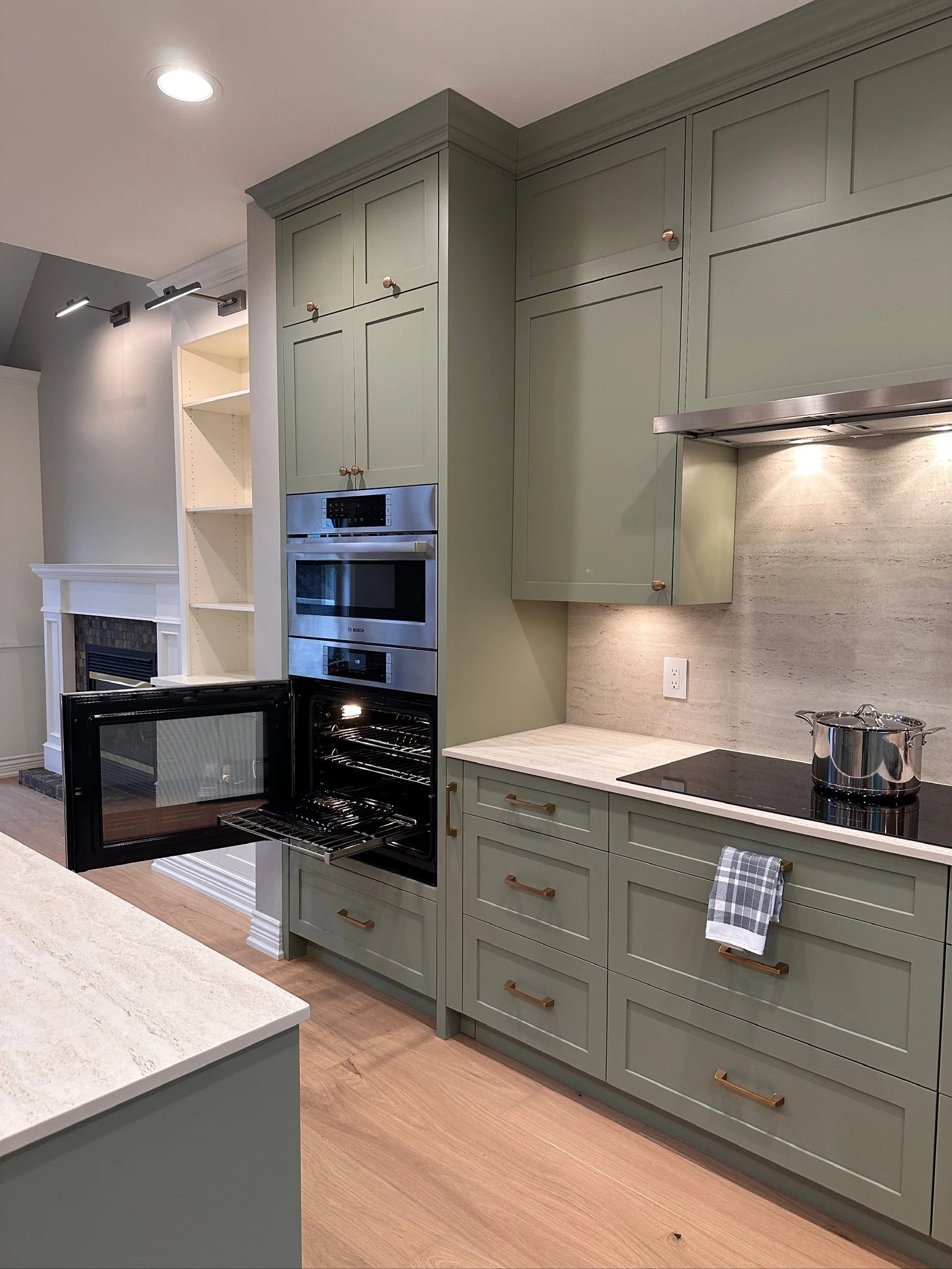 Kitchen with green cabinetry, double oven, black stove with pot, light wood flooring, and beige countertop with backsplash. Open oven door shows empty racks.
