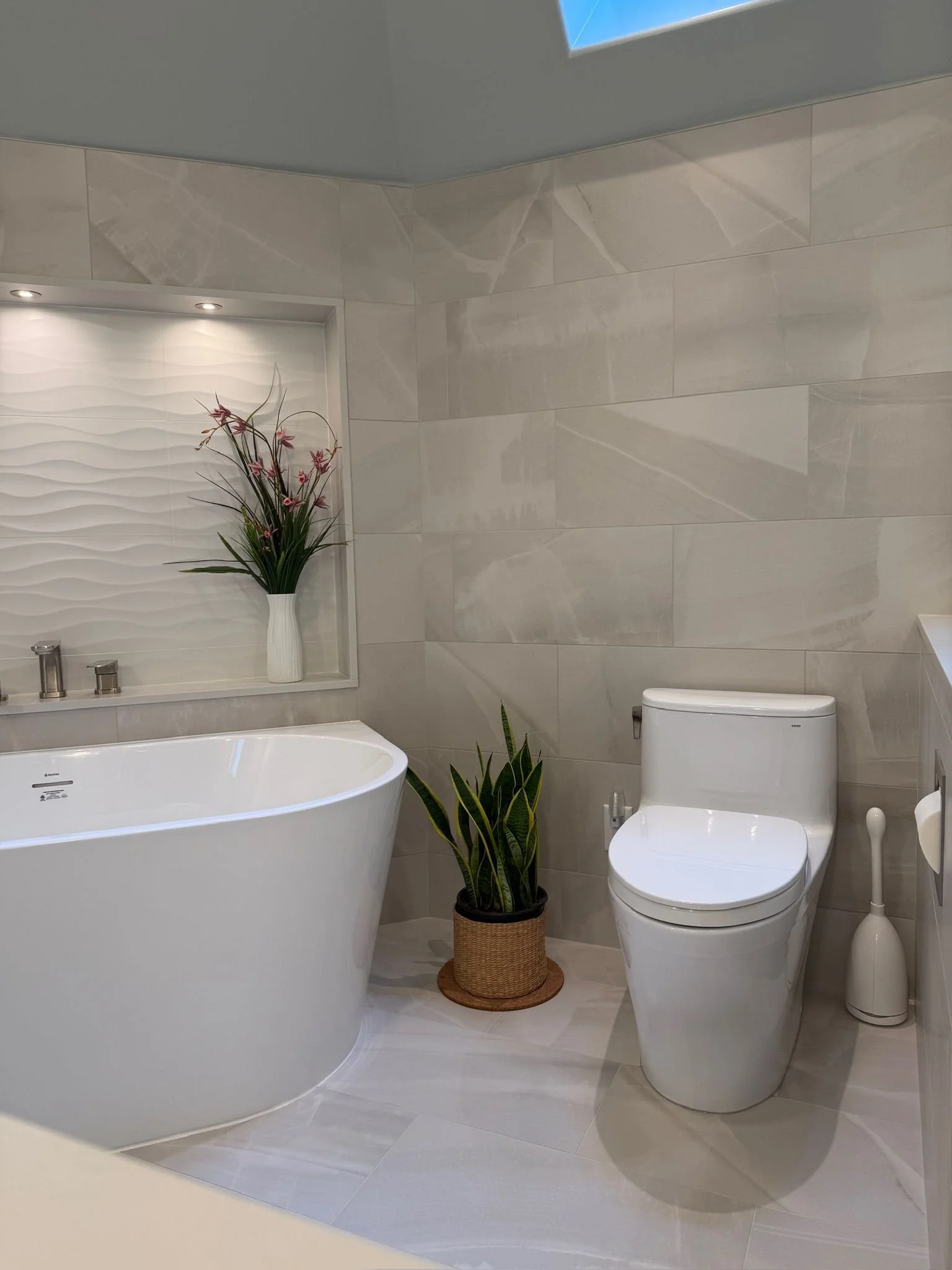 Modern bathroom with a white freestanding bathtub, a white toilet, potted plants including a tall leafy plant and a flowering plant in a woven basket, and decorative wall tile. Recessed ceiling light illuminates the space.