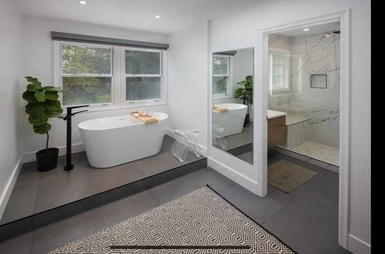 Modern bathroom with a freestanding bathtub, large windows, and a separate walk-in shower with marble tiles. Includes a mirror on the wall and potted plants for decoration.