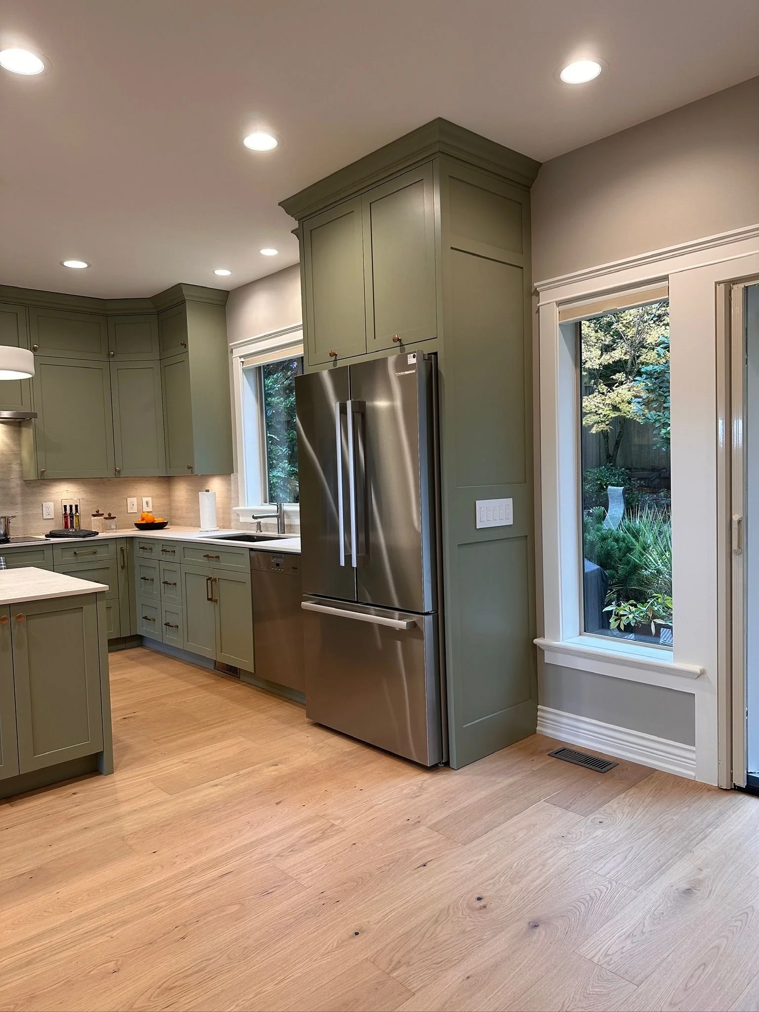 Kitchen with green cabinets, stainless steel refrigerator, light wood floor, and large windows showing outdoor greenery.