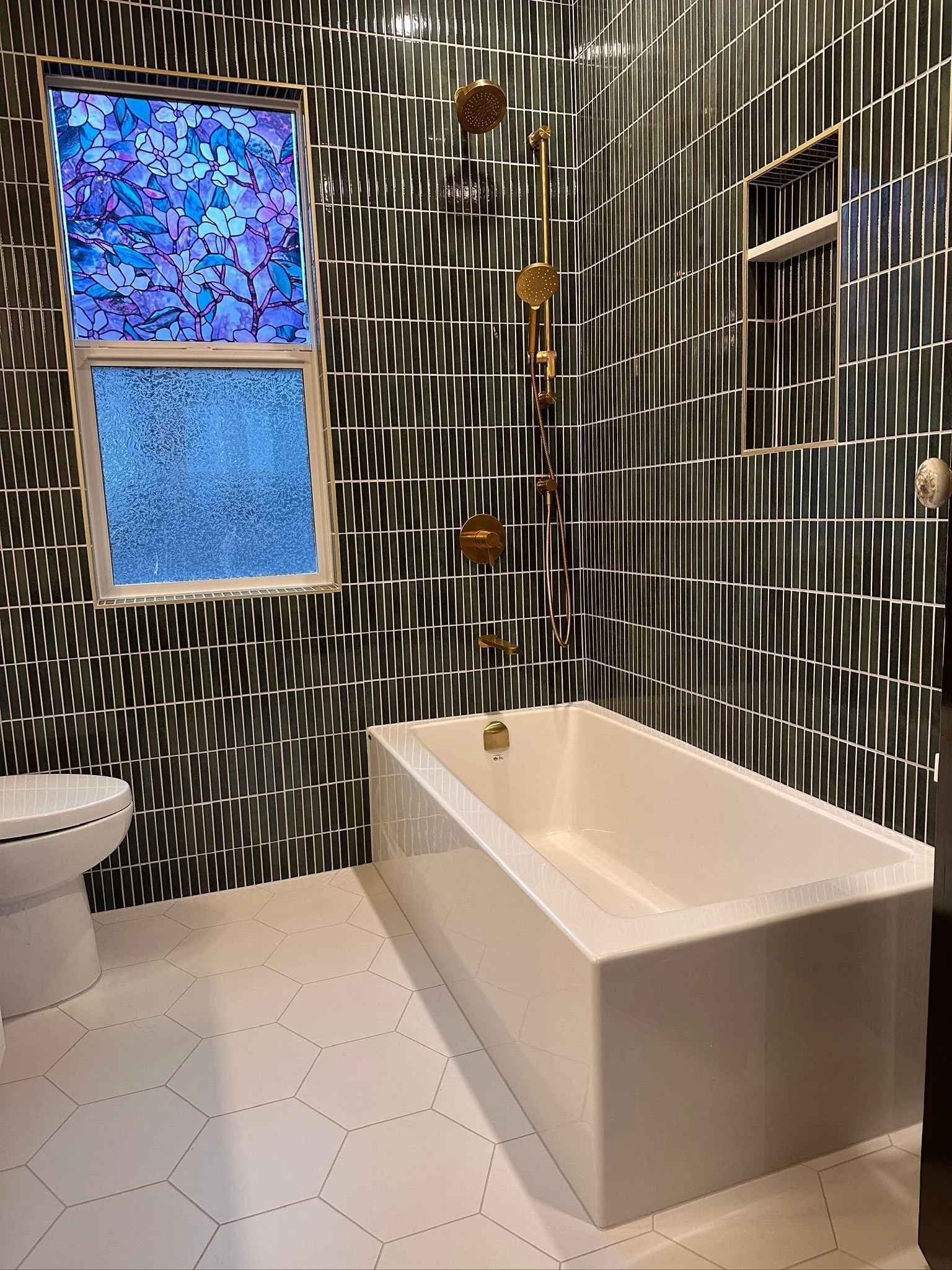 Modern bathroom with black rectangular tiled walls, white hexagonal tiled floor, a white bathtub, a frosted window with stained glass top, and a brass showerhead and fixtures.
