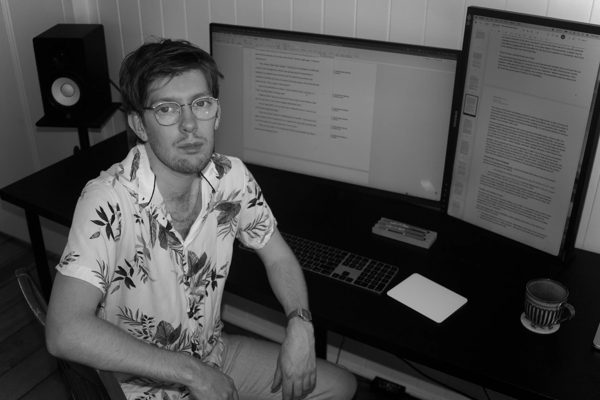 A young man with glasses and a patterned shirt sitting at a desk with two large computer monitors displaying documents, a keyboard, a mouse, and a cup on the desk.