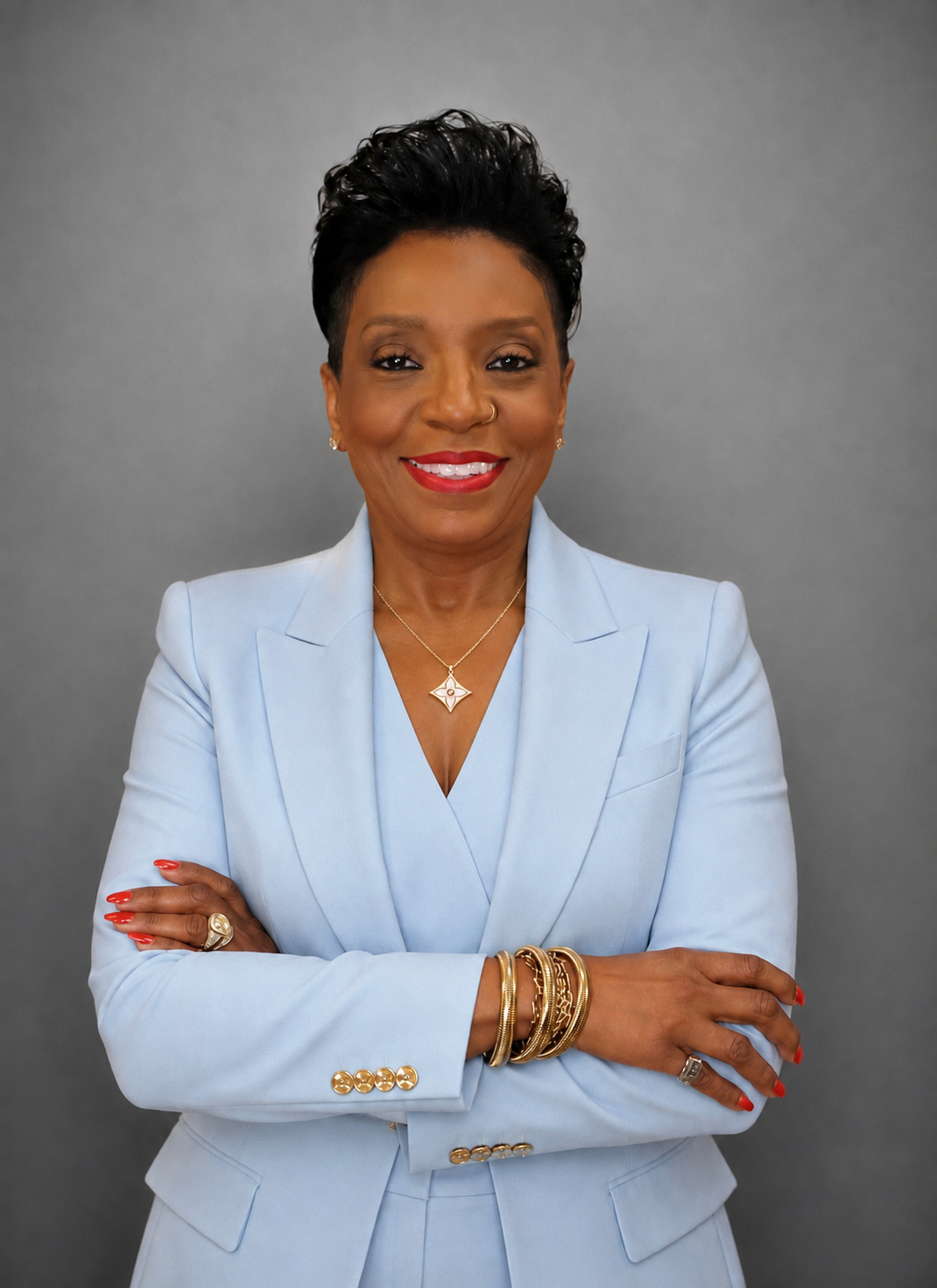 A confident African American woman with short black hair, wearing a light blue blazer and matching pants, standing with arms crossed, smiling, against a gray background.
