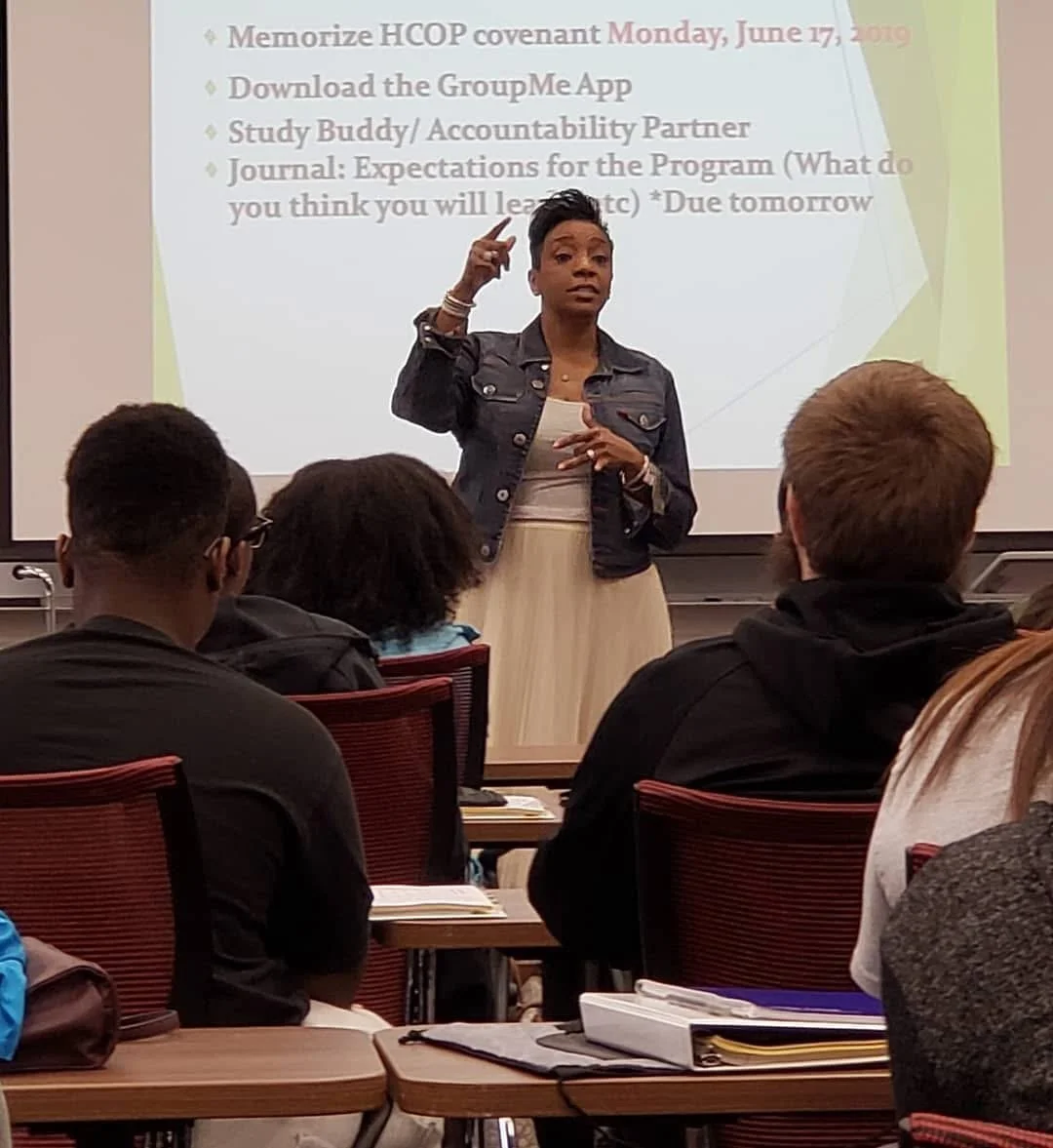 A woman stands at the front of a classroom, gesturing as she gives a presentation to students sitting at desks. A projected slide behind her lists tasks including memorizing a covenant, downloading an app, and studying with a partner.