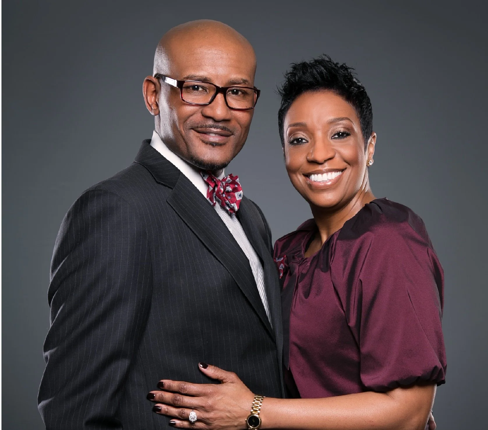 A man and woman in formal attire standing close together and smiling at the camera. The man is wearing a dark pinstripe suit with a white shirt, a patterned bow tie, and glasses. The woman is wearing a maroon satin blouse and has short black hair. She is smiling with her teeth showing and has her hand resting on the man's waist.