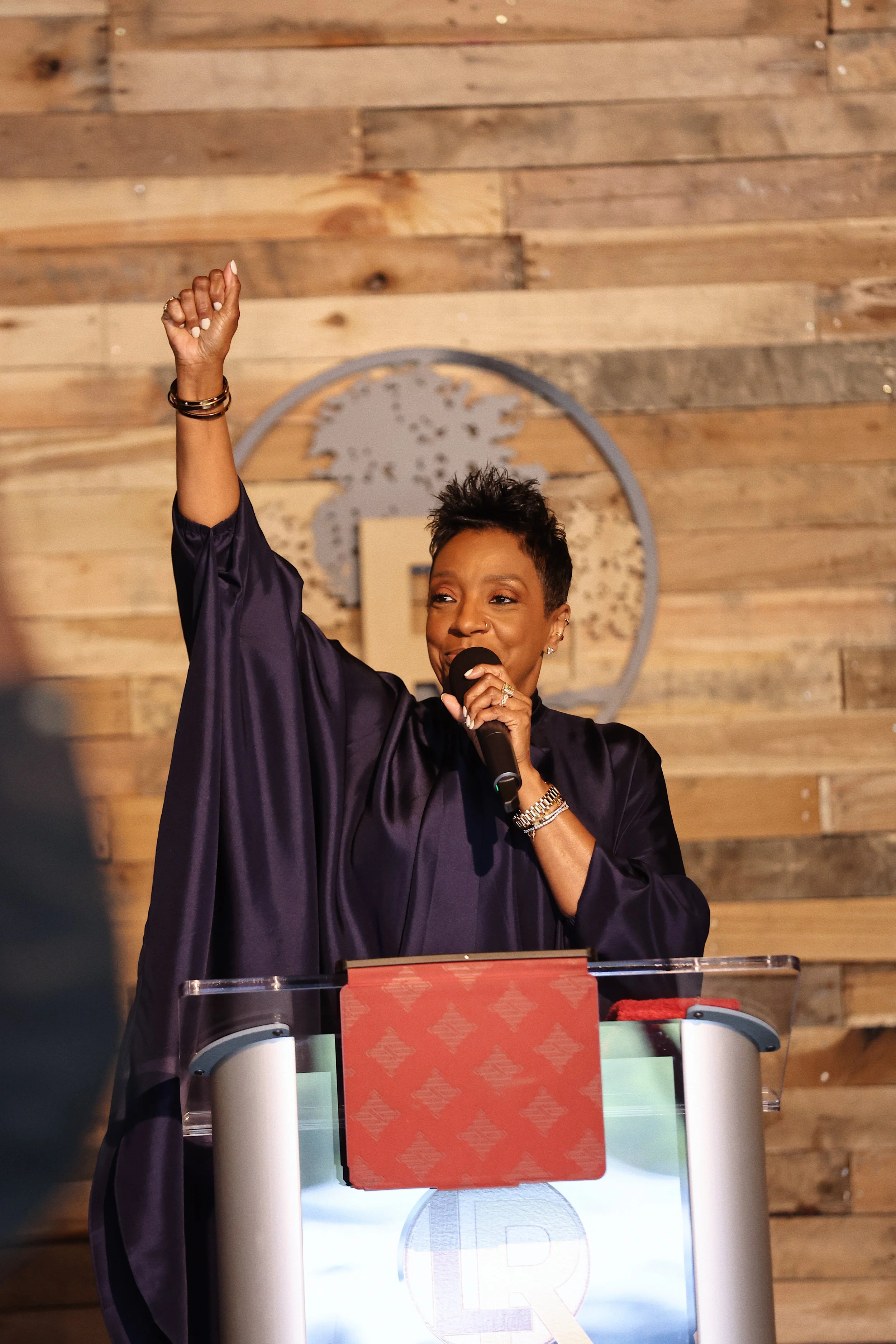 A woman at a podium speaking into a microphone with her right hand raised, wearing a dark purple robe, in a setting with a wooden wall and a circular logo behind her.