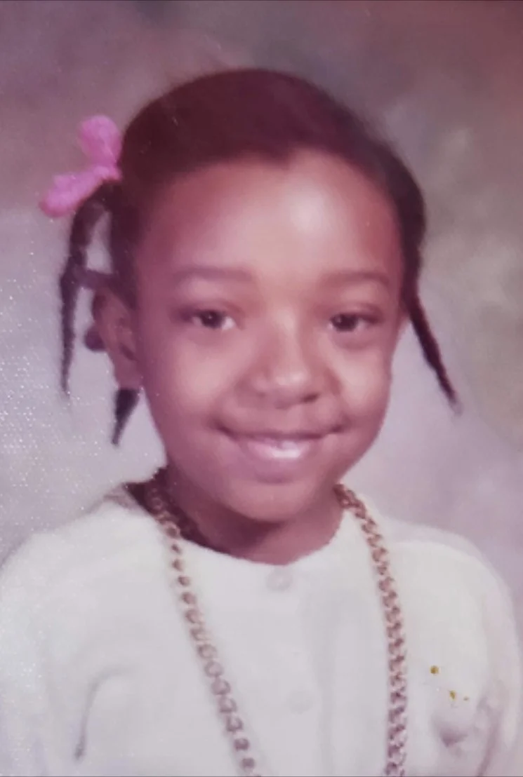A young girl with braided hair and a pink ribbon in her hair, wearing a white top and a gold chain necklace, smiling at the camera.