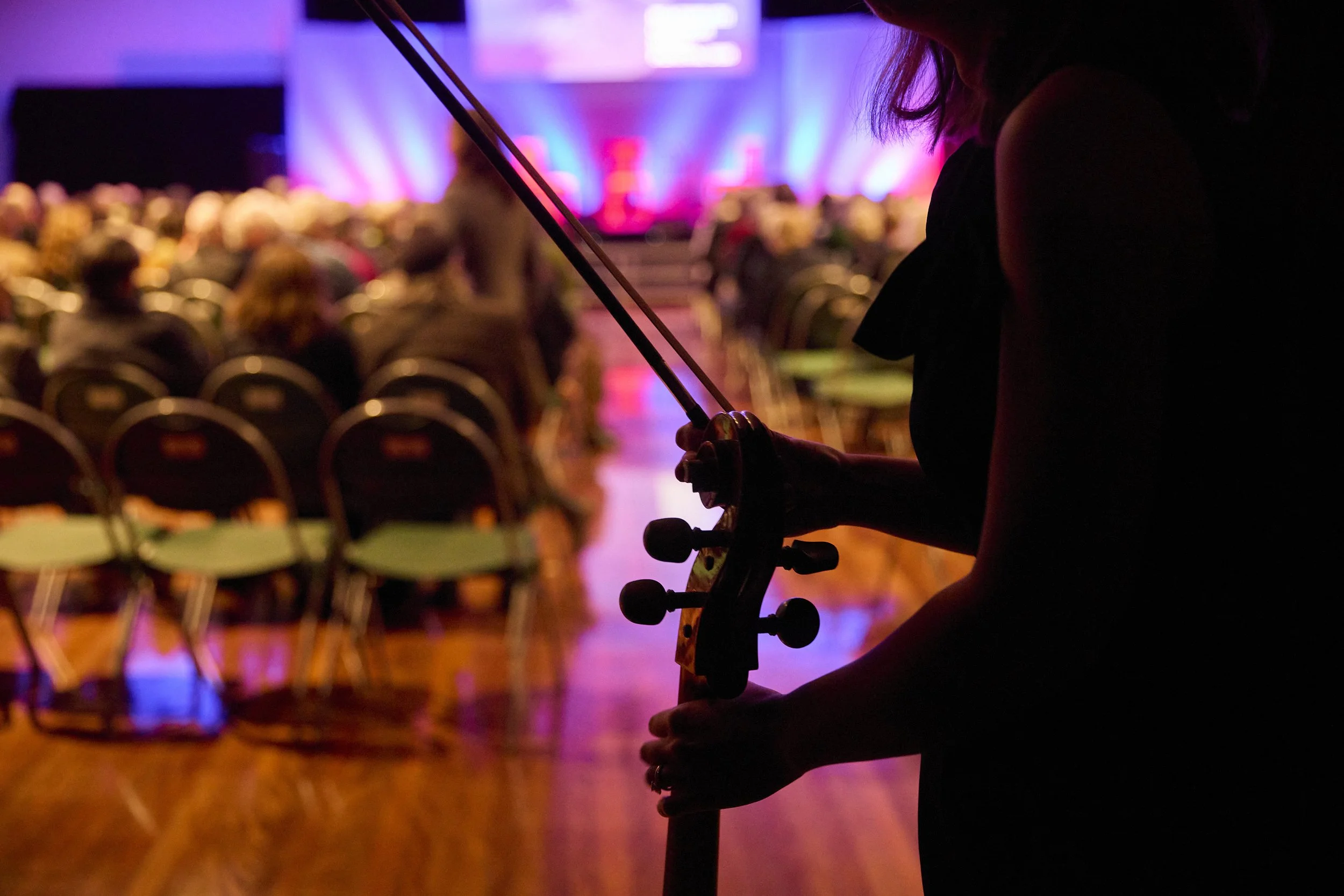 A person holding a cello in an event hall, with the audience and a stage in the background illuminated by colorful lights.