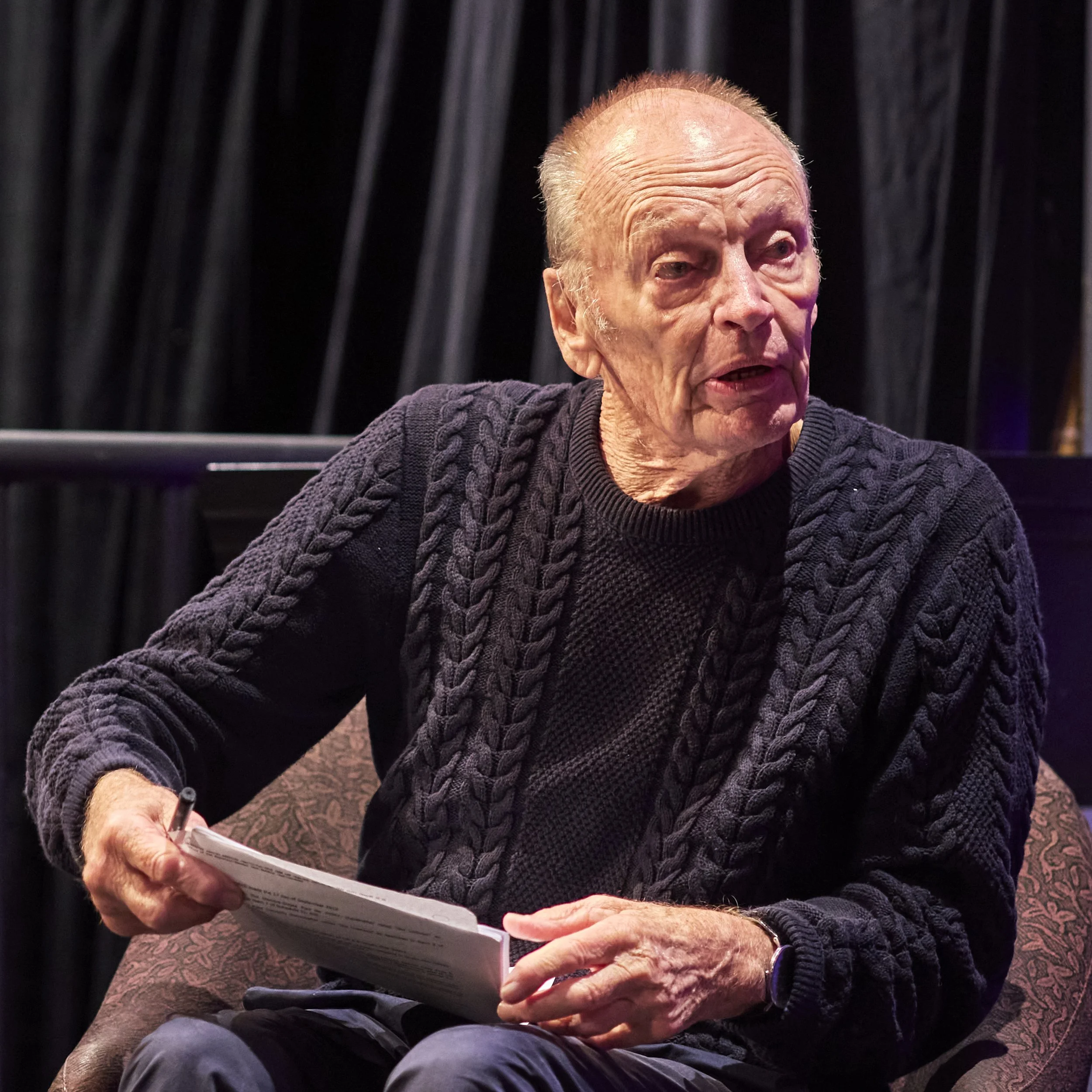 Close-up of John Bell, wearing a dark cable-knit sweater, holding a notes, sitting on stage with black curtains during a rehearsal.