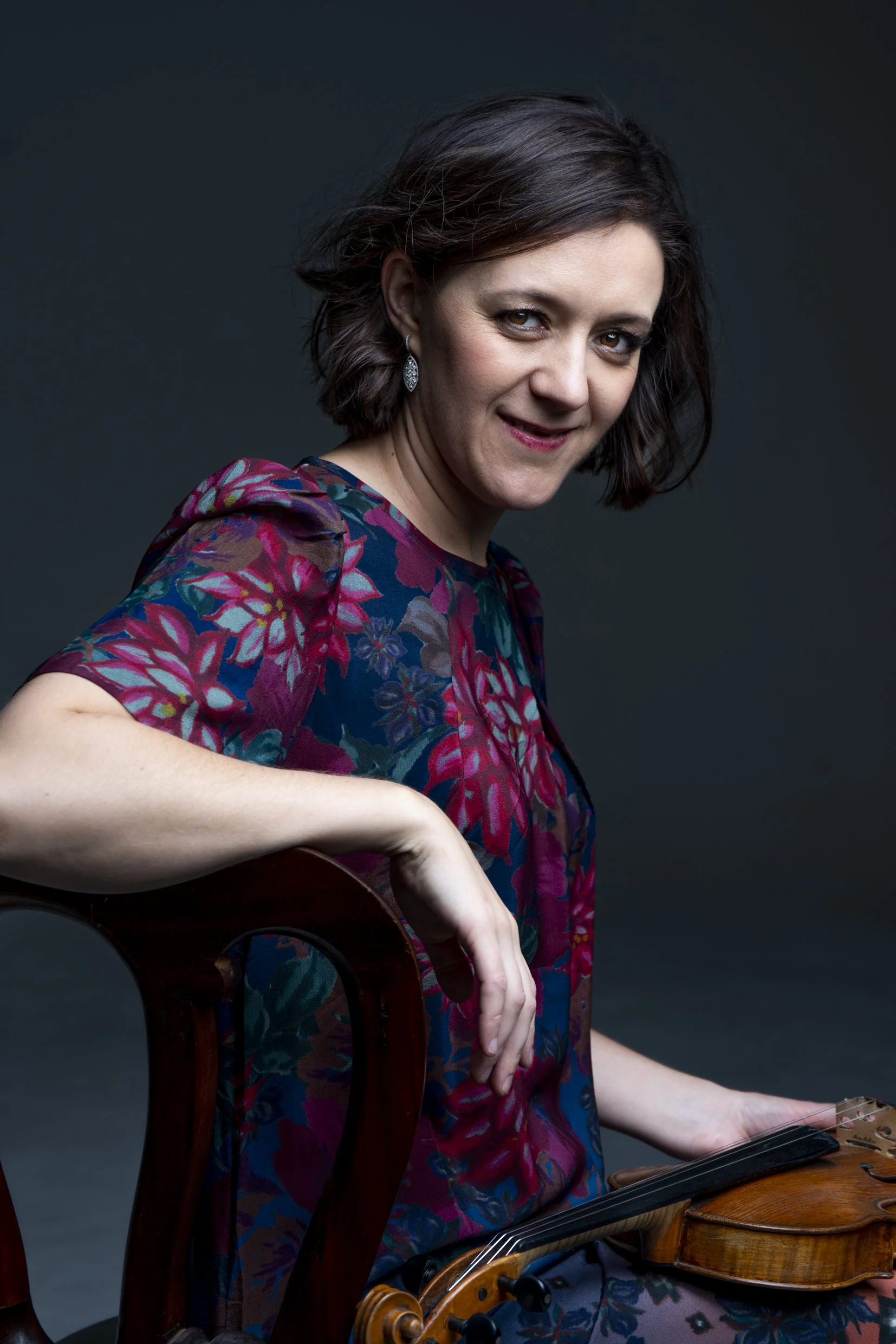 Lerida Delbridge with short brown hair wearing a floral dress, sitting on a wooden chair, holding a violin and a bow.