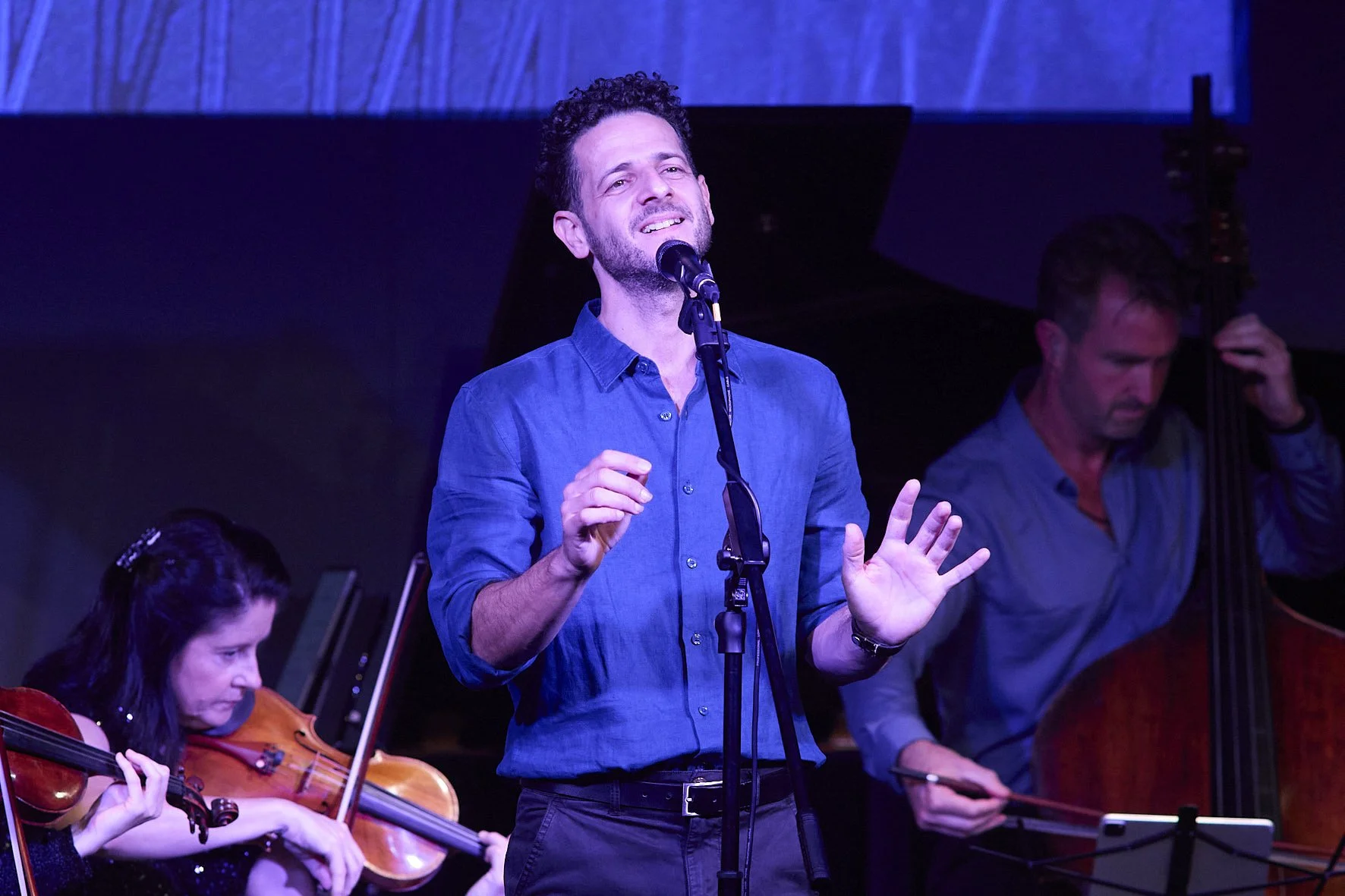 The singer Lior, singing passionately into a microphone on stage, with musicians playing string instruments next to him, during a live performance.