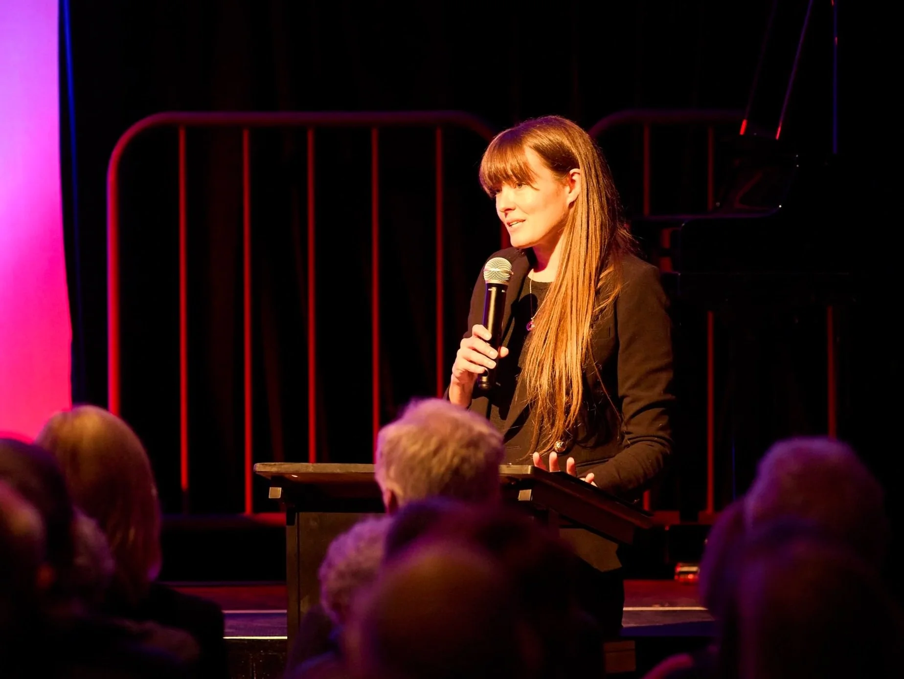Alice Coffey with long hair holding a microphone in speaking at a podium as MC before a performance or presentation on a stage, with an audience watching.