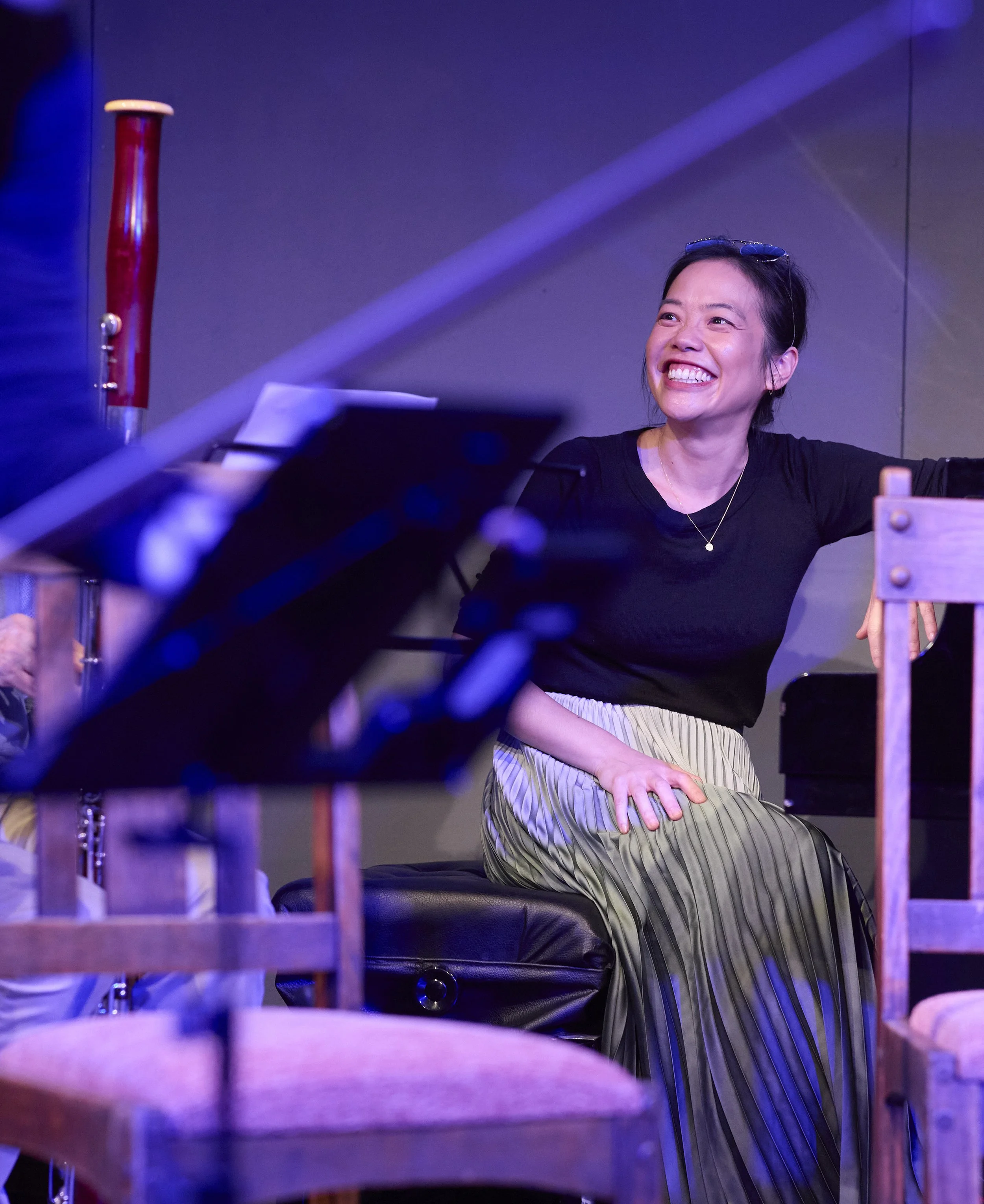 Pianist Andrea Lam sitting at a piano, smiling on stage during a rehearsal, with purple and blue lighting.