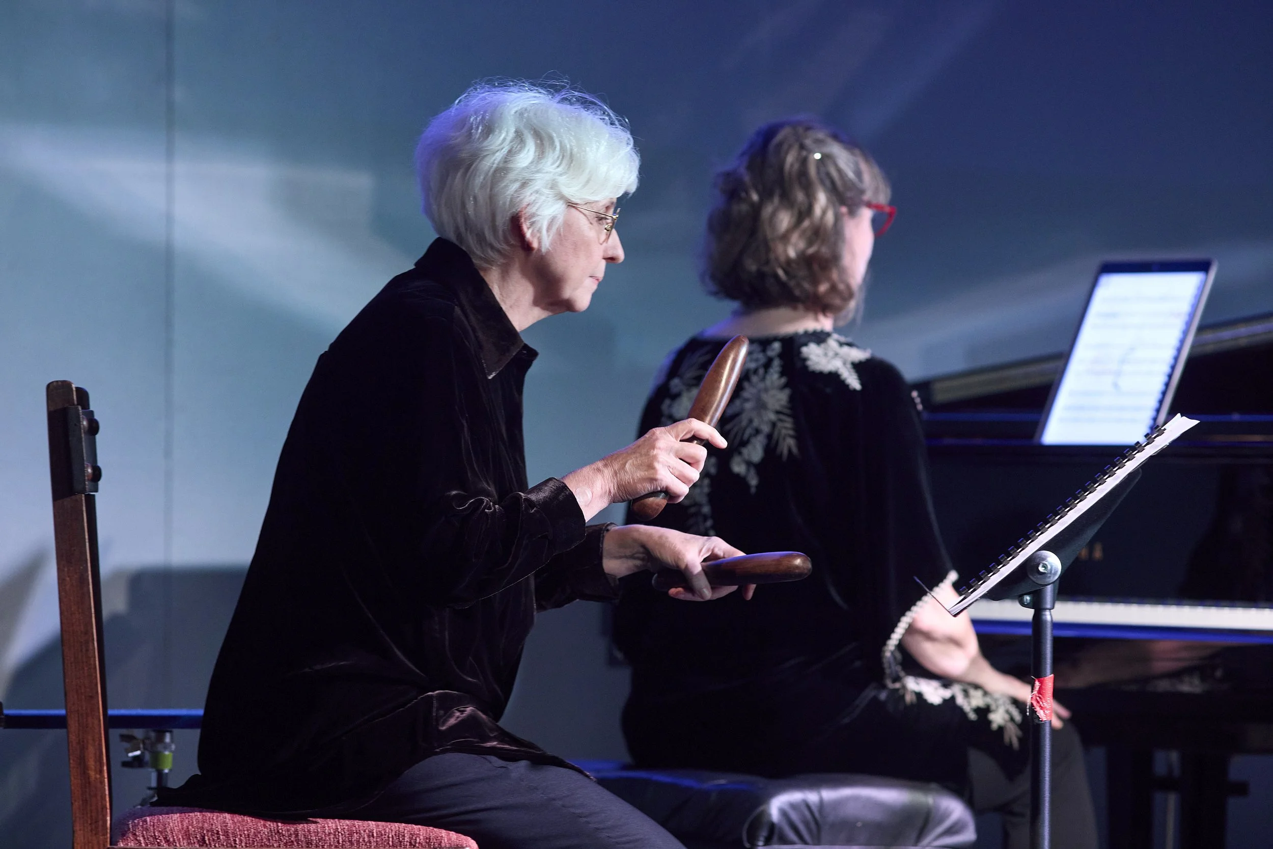 Percussionist Rebecca Lagos seated on a red chair playing claves,  with Bernadette Harvey playing the piano in the background.