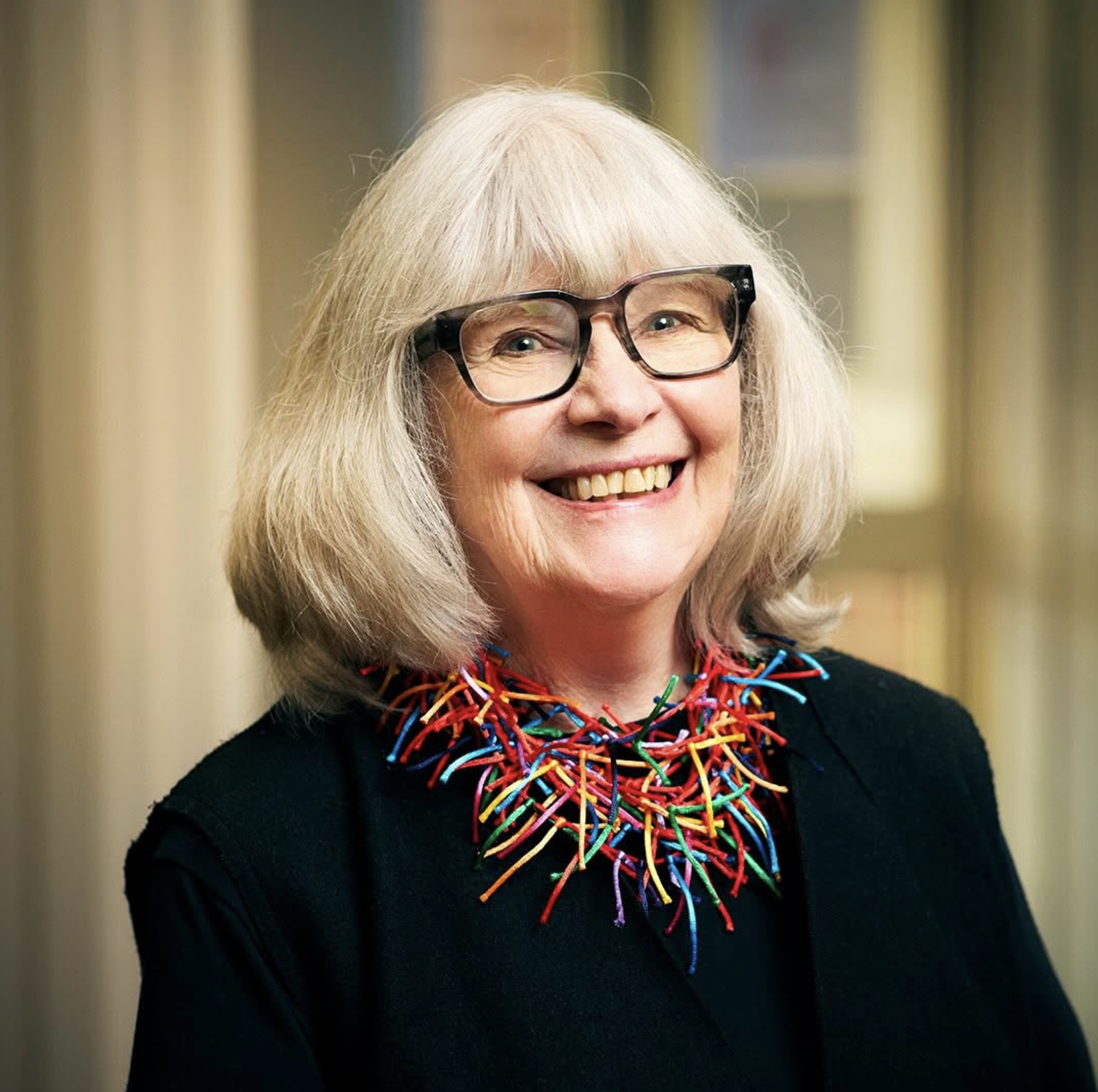 A smiling Catherine Harker, founder of BCMF, wearing glasses and a colorful necklace, against a blurred background.