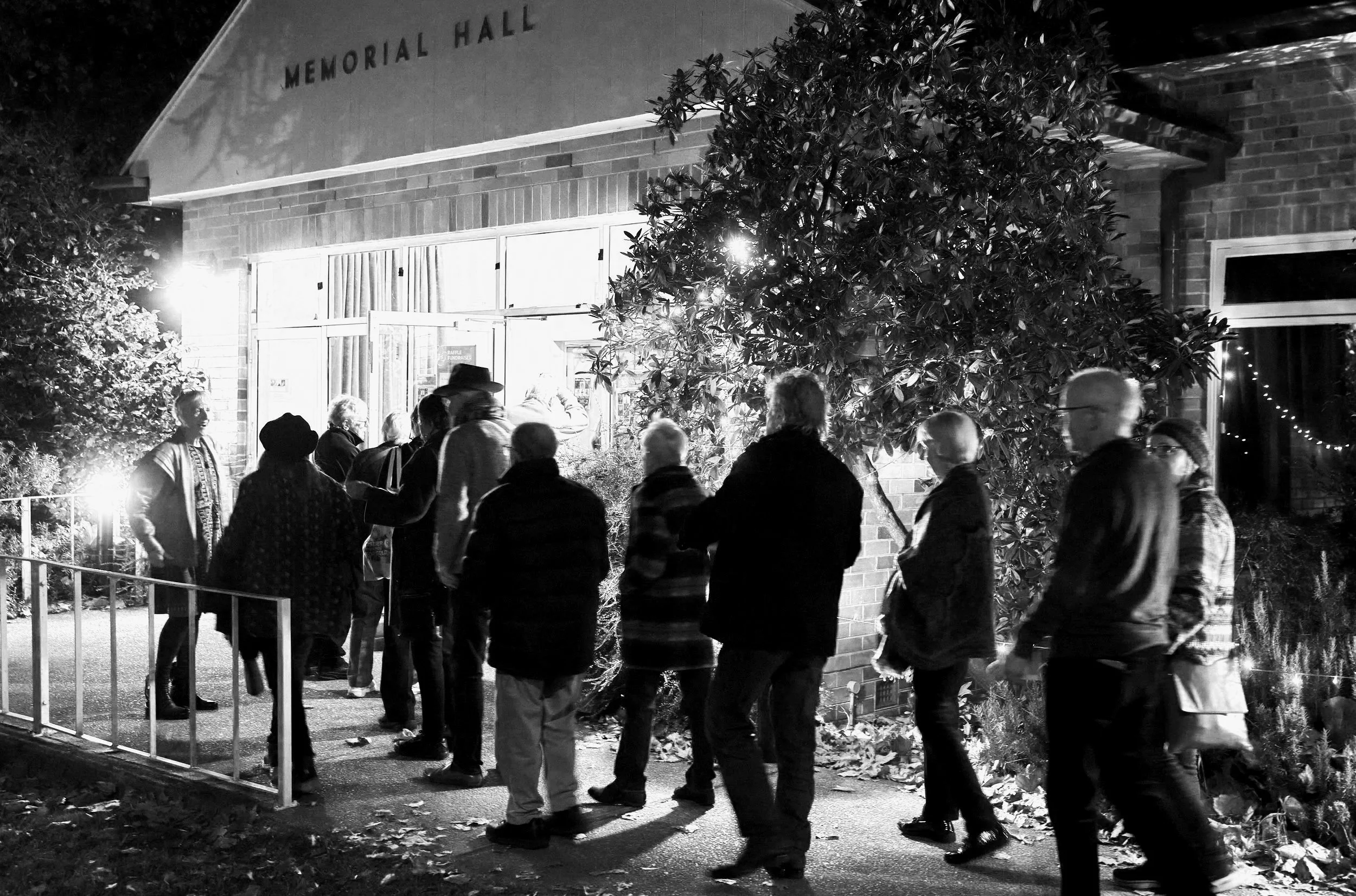 A black and white photo of a group of people standing outside the Phillips Hall at night. Some are wearing jackets and hats, and they are gathered near a tree and a railing. The Memorial Hall sign is visible on the building.