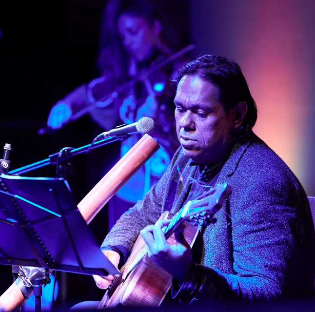 William Barton playing an acoustic guitar while Verronique Serret is in the background playing a violin on stage, with purple and blue lighting.