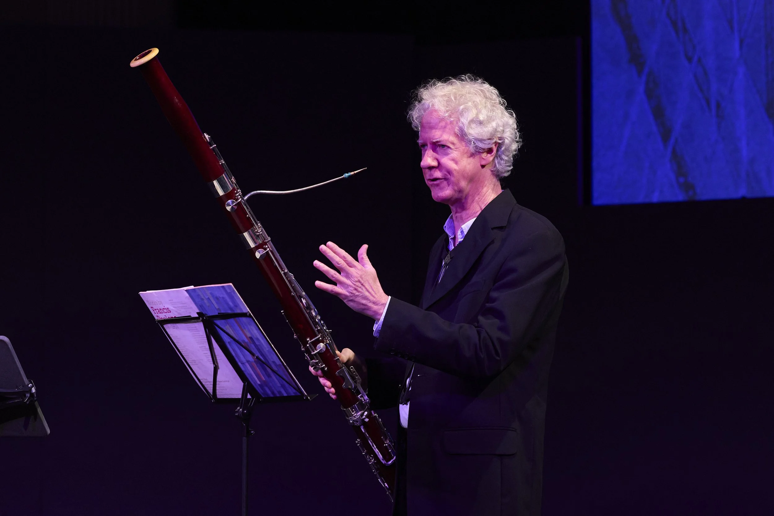 Matthew Wilkie playing a bassoon on stage during a performance, with music sheets on a stand in front of him and a purple-lit screen in the background.