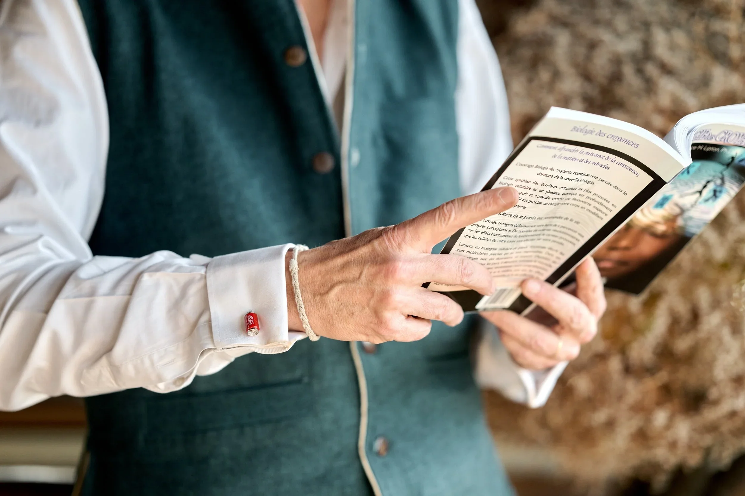 A person in a dark green vest and white shirt is reading a book, with a gold bracelet on their right wrist and a small red and white Coca-Cola-branded wristband on their left wrist.