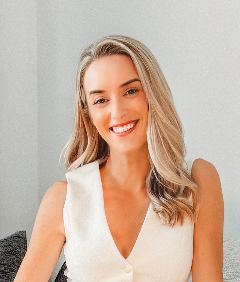 A smiling woman with blonde hair wearing a white sleeveless top, sitting on a gray couch in a light-colored room.