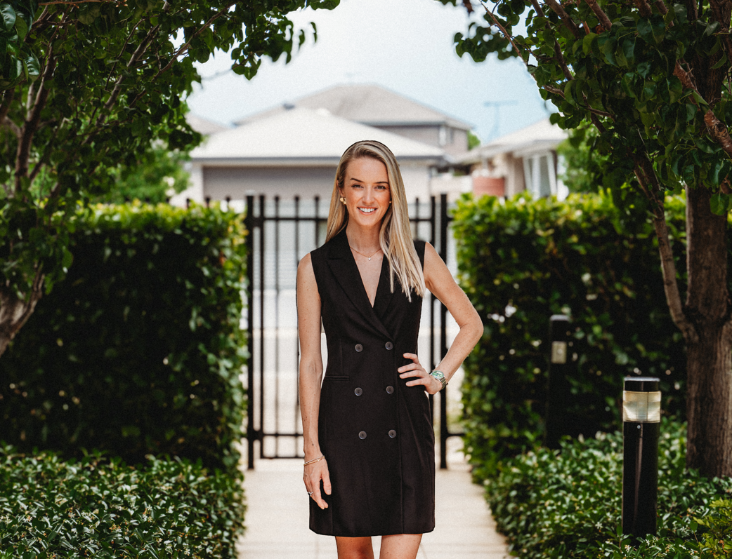 A woman with long blonde hair wearing a sleeveless black dress standing on a garden pathway, smiling, with bushes, trees, and residential houses in the background.