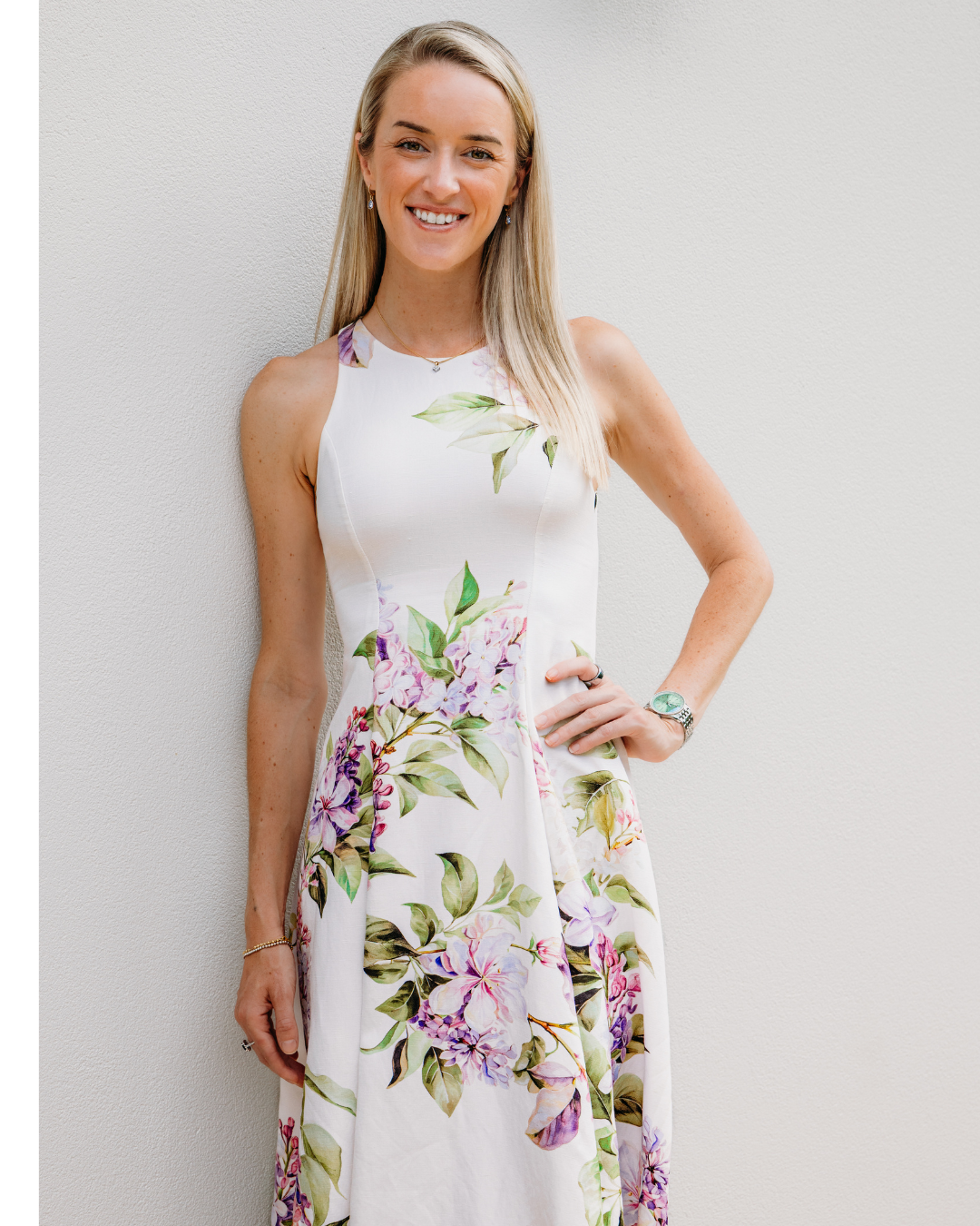 A smiling woman with long blonde hair wearing a white floral dress, standing against a plain wall.