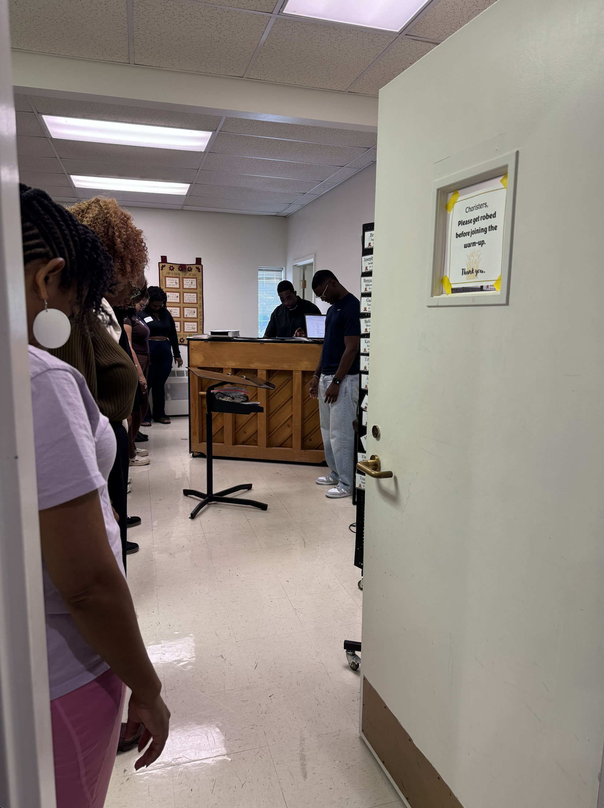 View of people standing in a room with ceiling lights, a piano, and a sign on the door that requests robed choristers to get ready before warming up.